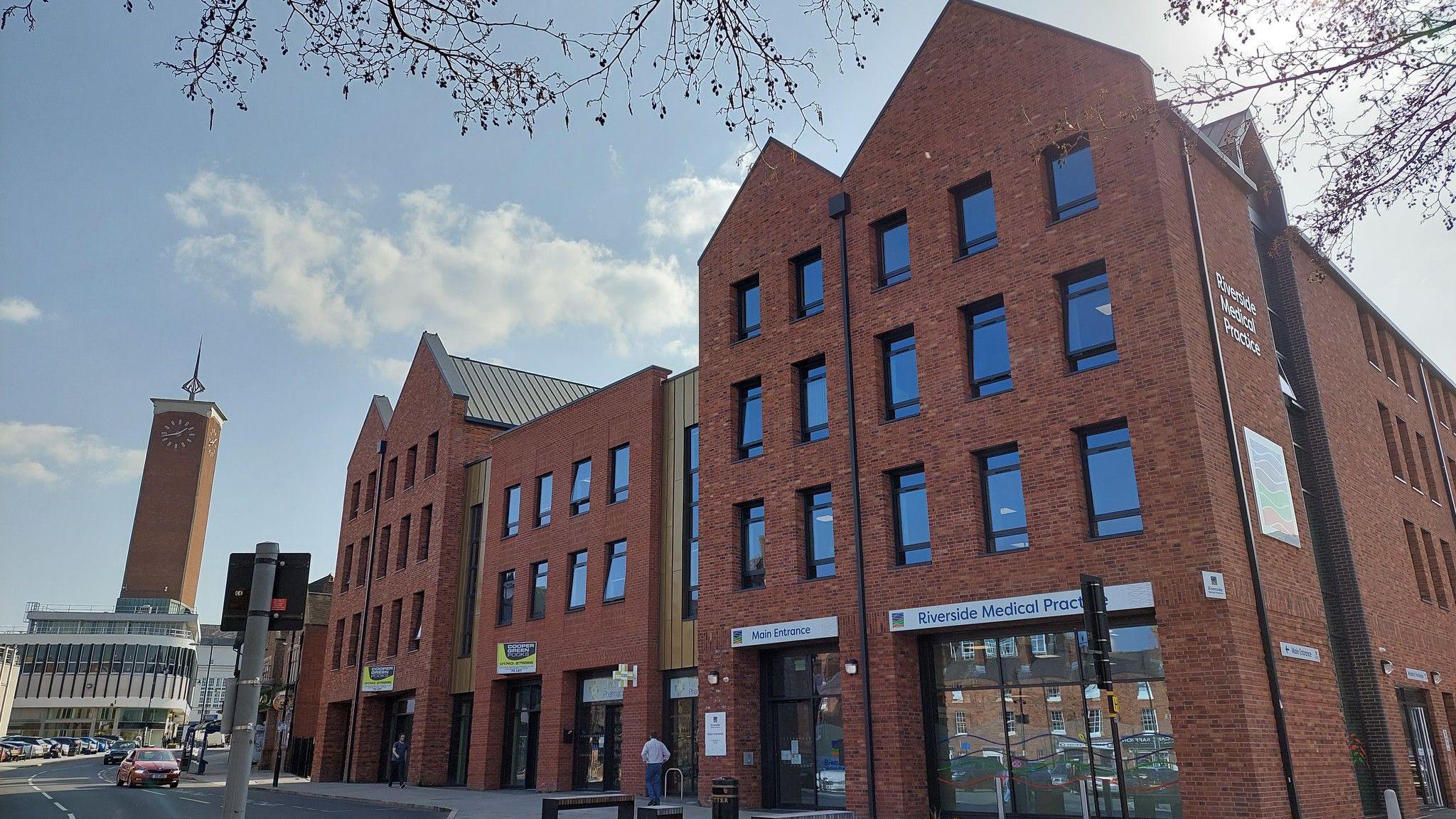 A large modern red-brick multi-storey building with a sign for Riverside Medical Practice above the ground floor window. A large clock tower can be seen to the left, with cars on the street.