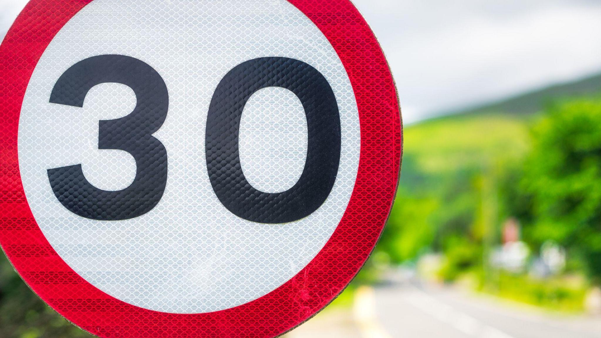 A close up of a 30mph road sign on a sunny day, with trees in soft focus behind it.