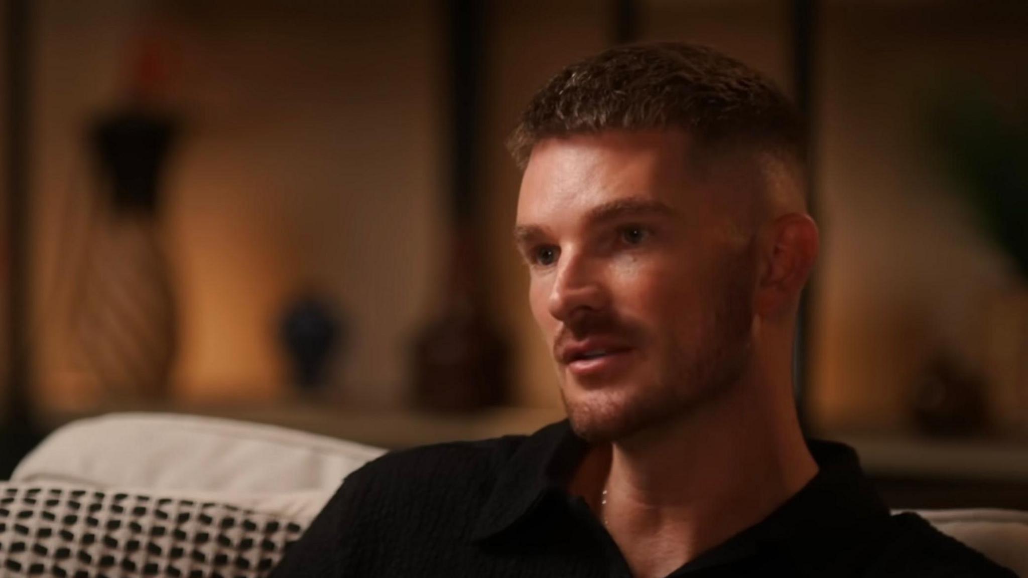 Adam Frisby, a white man with short, fair hair and a short beard. He is sitting on a sofa being interviewed for a podcast