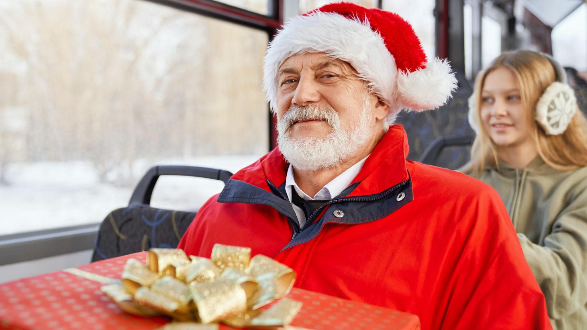Old man in santa hat smiling holding present and girl sitting behind him in moving bus.