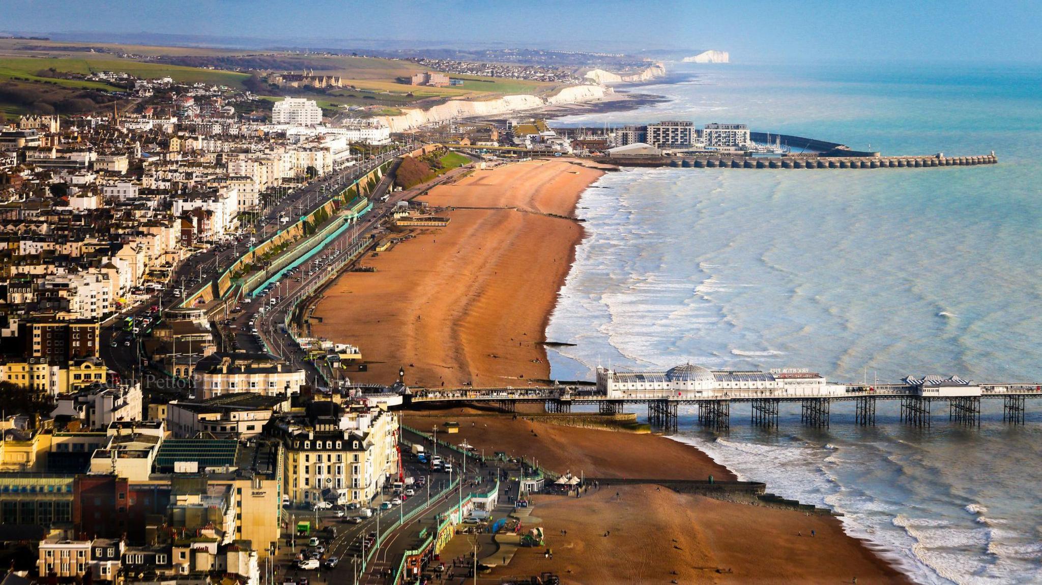 A wide-angle aerial view of the beach and coastal town of Brighton, with two piers extending out into the sea and white cliffs in the distance. 