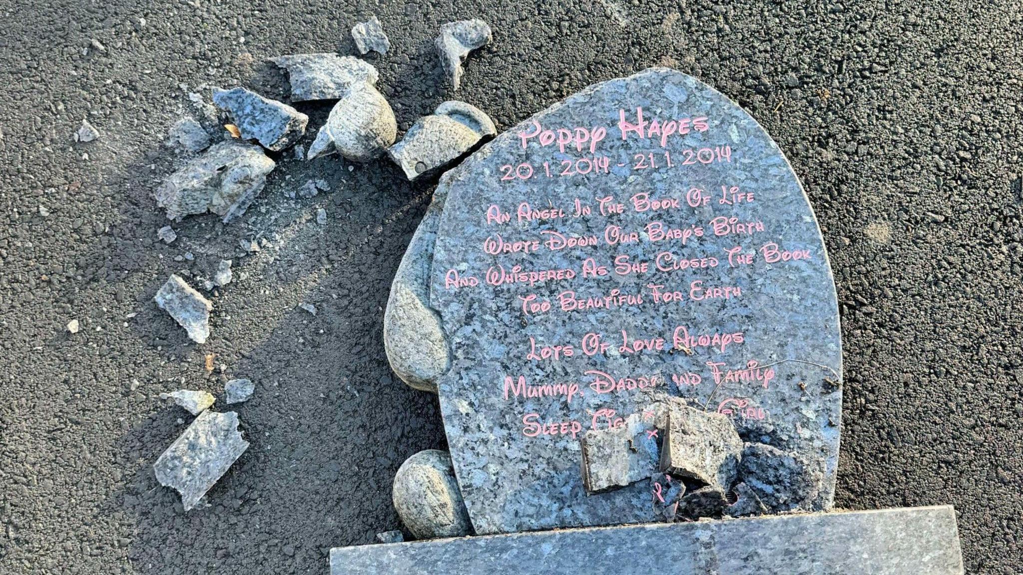 A photograph of the damaged gravestone. It is on the floor with bits of stone broken off the top of it. 