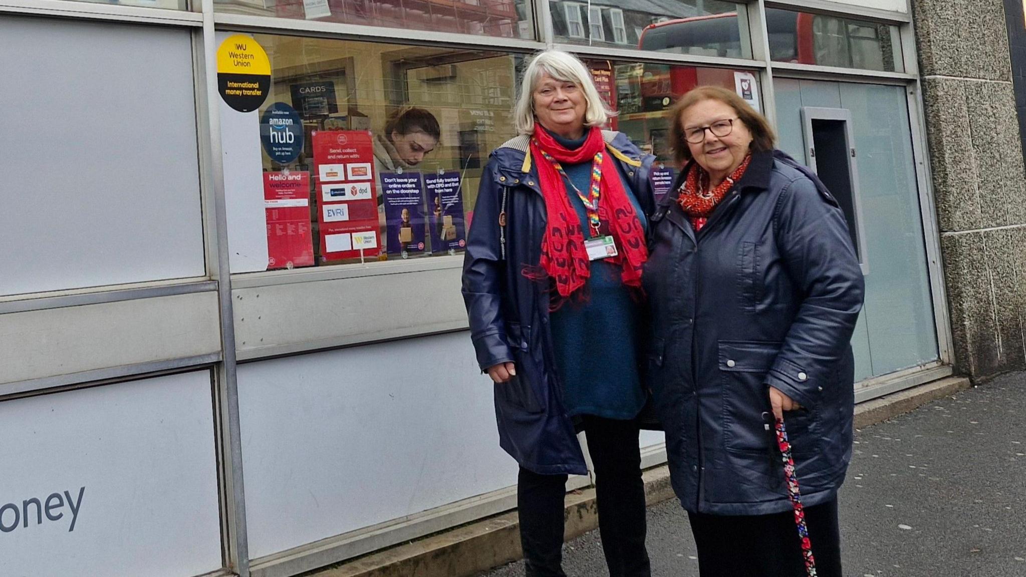 Councillors Angela Penrose and Mary Aspinall outside Mutley Plain Post Office. The pair are wearing navy coats and black trousers. They are looking at the camera and smiling. Ms Penrose has light hair and is wearing a red scarf. Ms Aspinall has brown hair. 