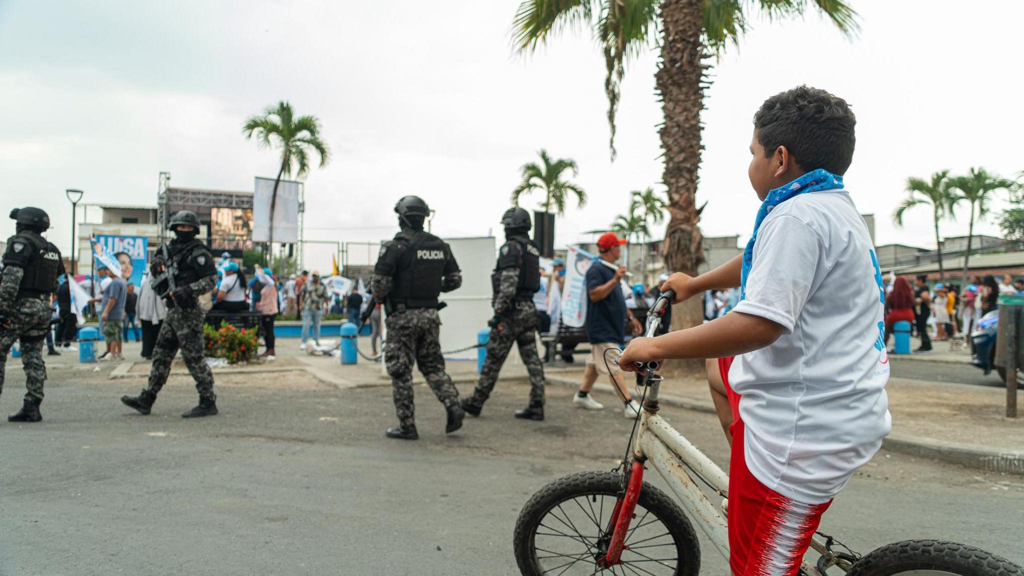 A boy on a bicycle watches as armed police patrol