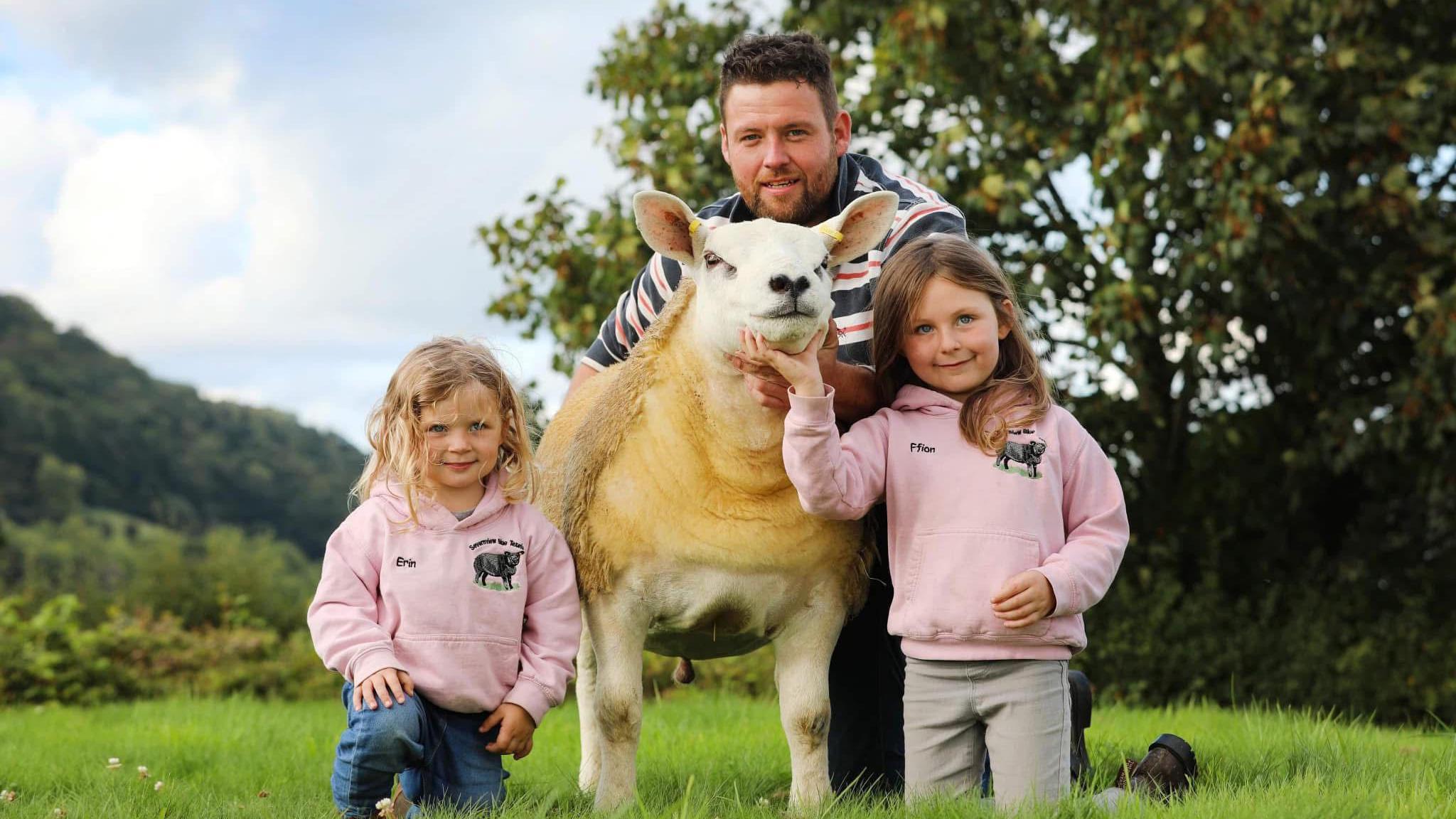Michael Davies kneels behind a sheep. He is wearing a black and red and white stripey jumper and has short brown hair and stubble.One of his young daughters, who looks about five, kneels next to the sheep in a pink jumper and blue jeans. She has curly blonde hair. On the right of the sheep Davies' older daughter, who looks to be about seven or eight wears a matching pink hoodie and grey jeans. She has long brown hair. She is holding the sheep's head. Bind them in the background a tree and bushes are visible. 