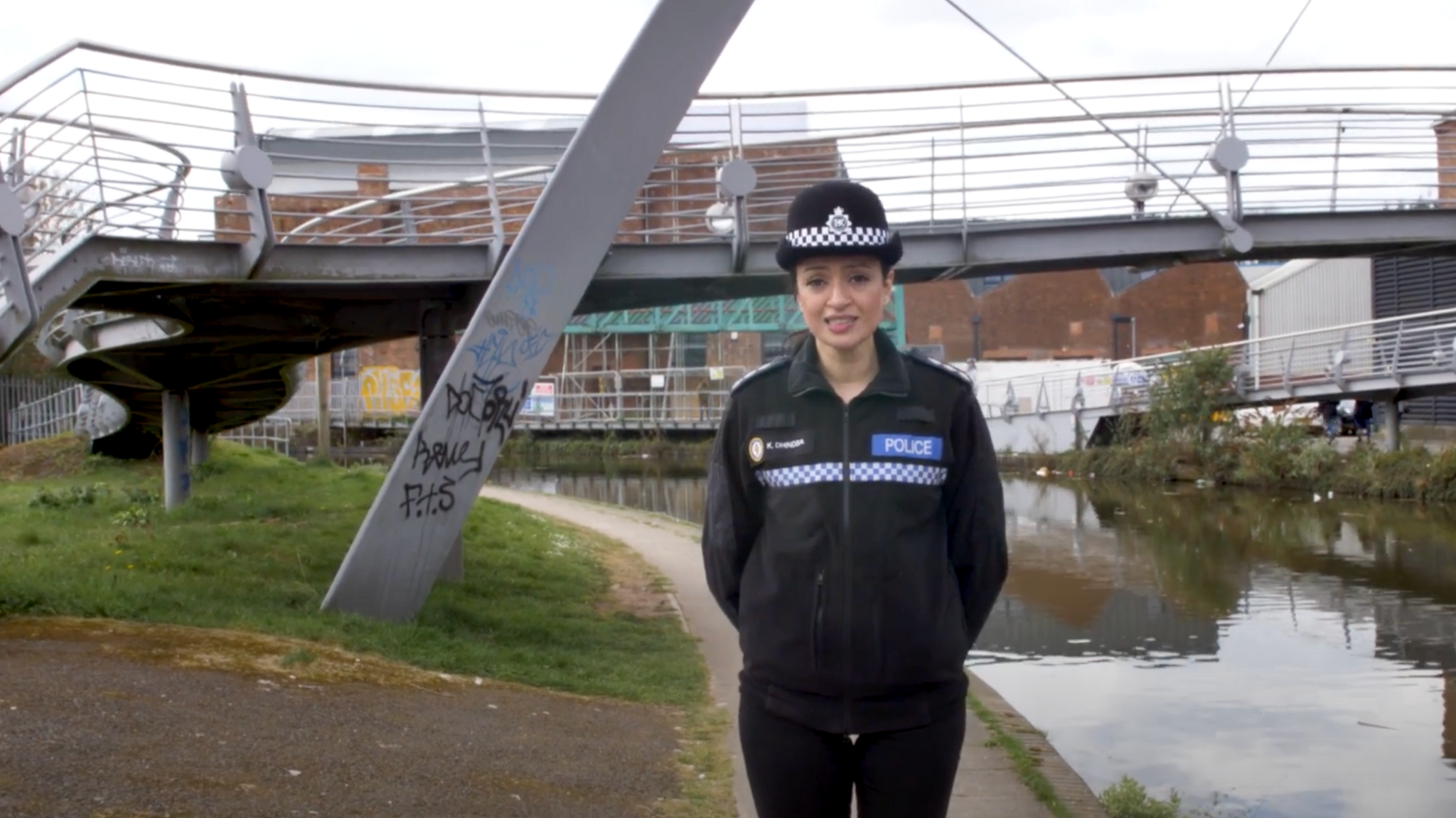 Khizra Bano standing next to a canal in Coventry, wearing her police uniform. A metal footbridge crosses the canal behind her, a grey structure with thin metal railings. One of the supporting spars has graffiti on the side.