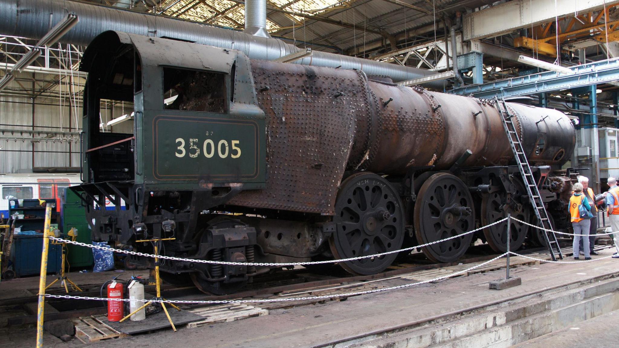 A rusting section of the locomotive is up on a platfrom inside a warehouse, it is behind a fenced of section, there is a ladder on the side of it and there is a long barrel which looks like it's rusting and a forest green coloured front section which has 35005 on the front