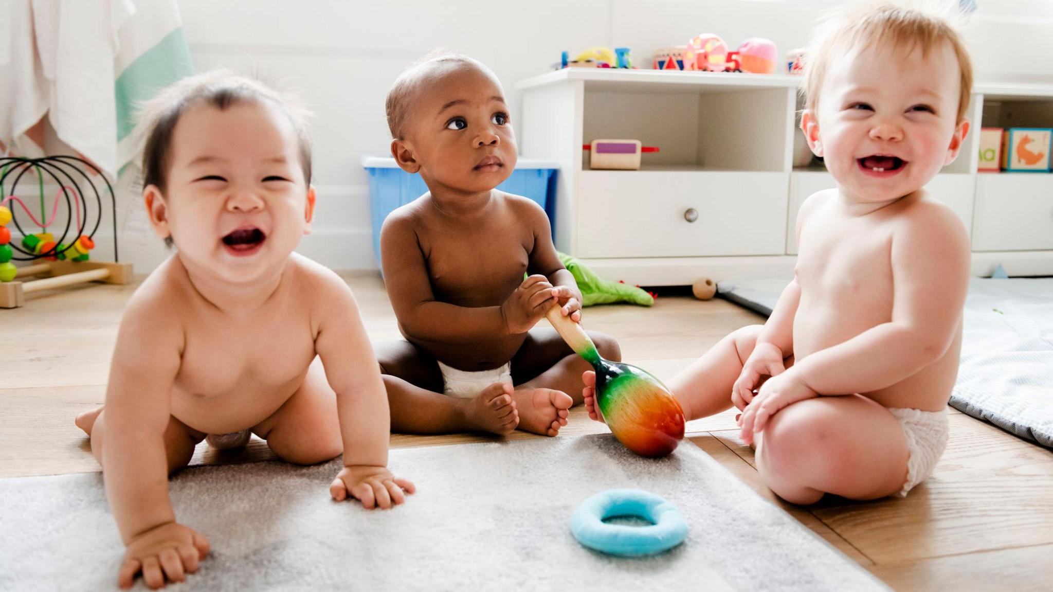 Three babies wearing diapers sit on the floor of a playroom, two laughing and one looking into the distance and holding a toy