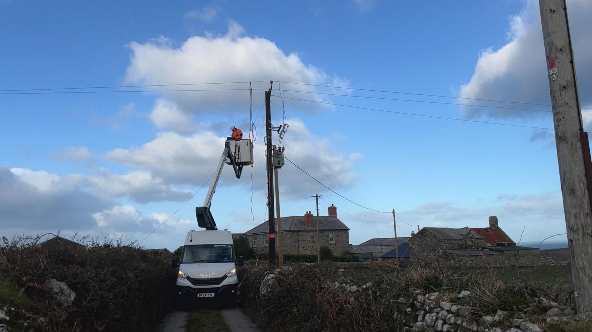 A white van in the middle of a country lane, with some houses in the background and overhead power cables. A person in orange overalls and a white hard hat in a cage works on the power cables.