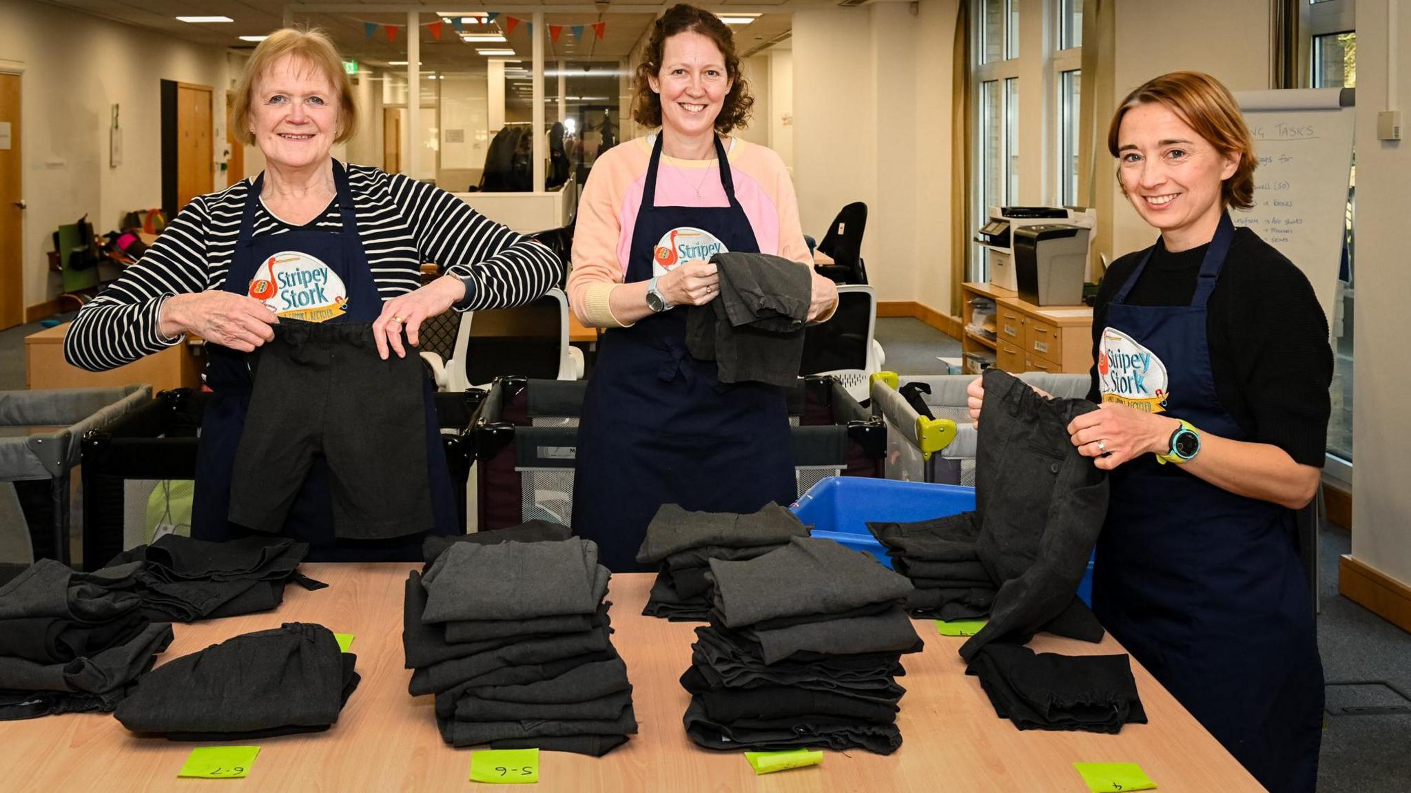 Three workers at Stripey Stork folding uniform items to be administered to children in Surrey. The three women are all wearing navy aprons with the charity's logo, and are all stood beside piles of grey uniform trousers. The lady on the left has blondish hair and is wearing a striped top. The lady in the middle has brown hair and is wearing a pink top. The lady on the right has light brown hair and is wearing a black jumper.