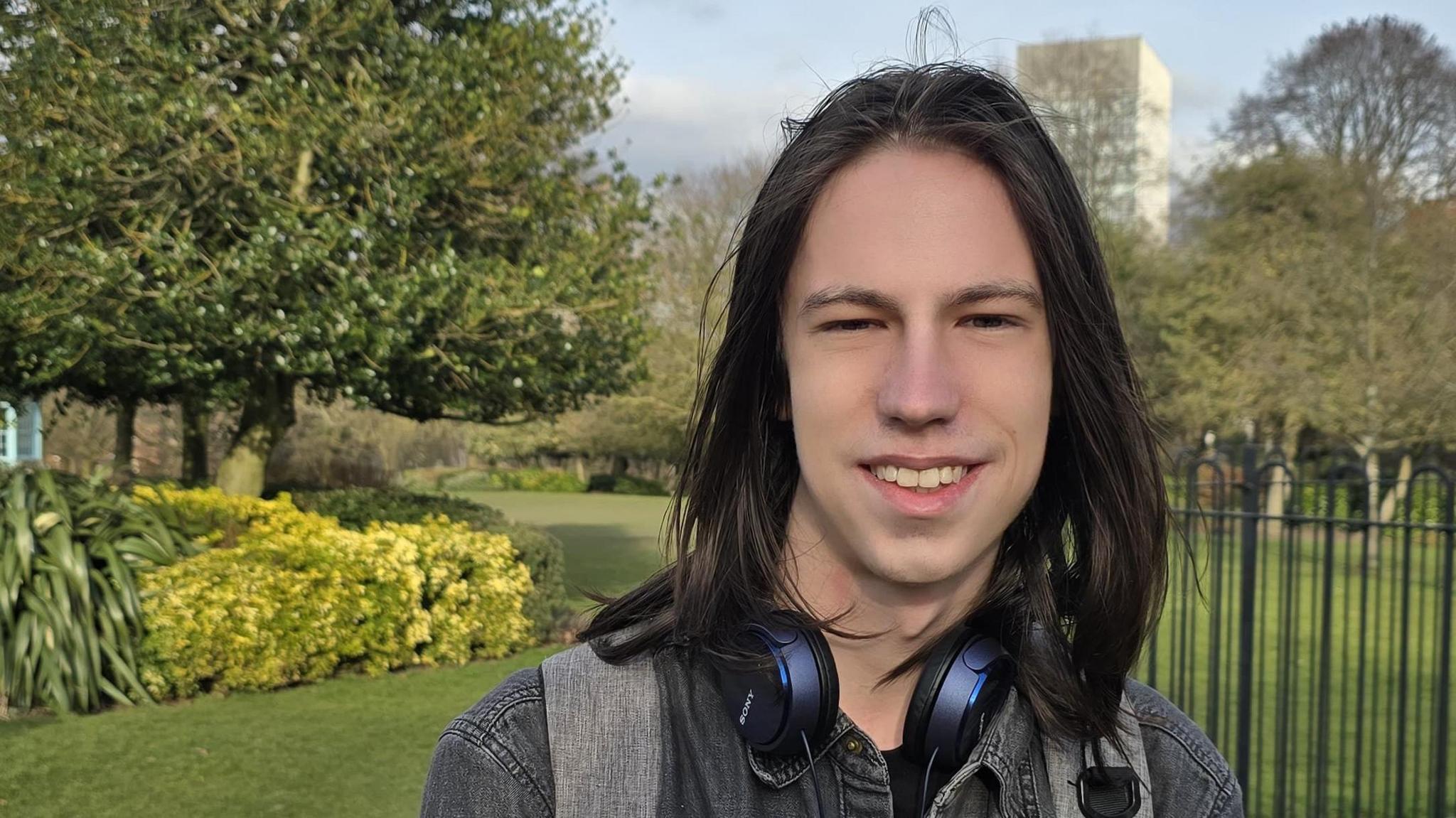 A young man smiles into the camera. He is clean shaven and has long dark hair down to his shoulders. He has headphones around his neck and is wearing a great denim jacket. He is stood in a park with a Sheffield University building in the background.