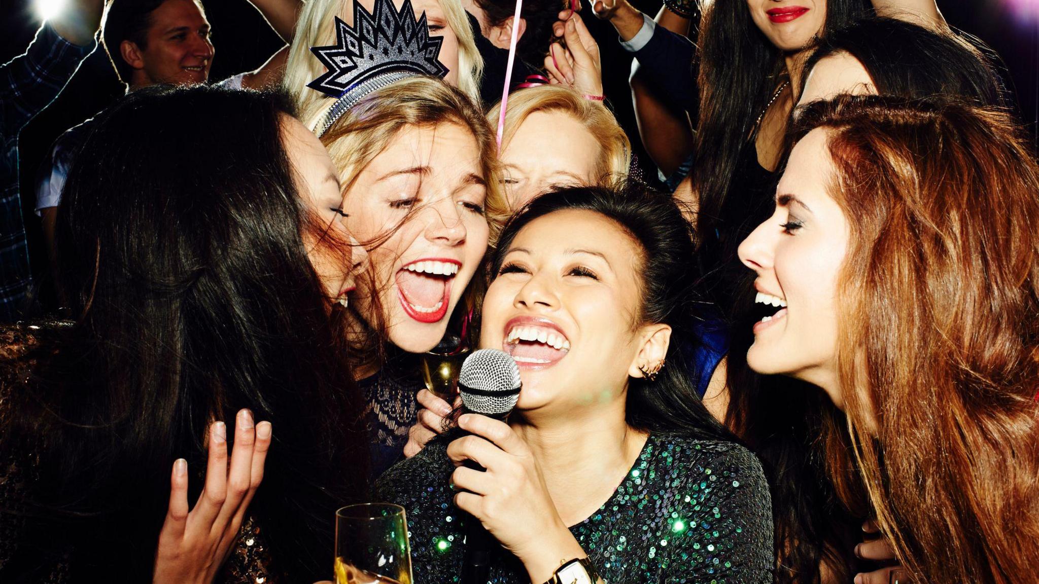 A group of women singing karaoke together, smiling and holding drinks