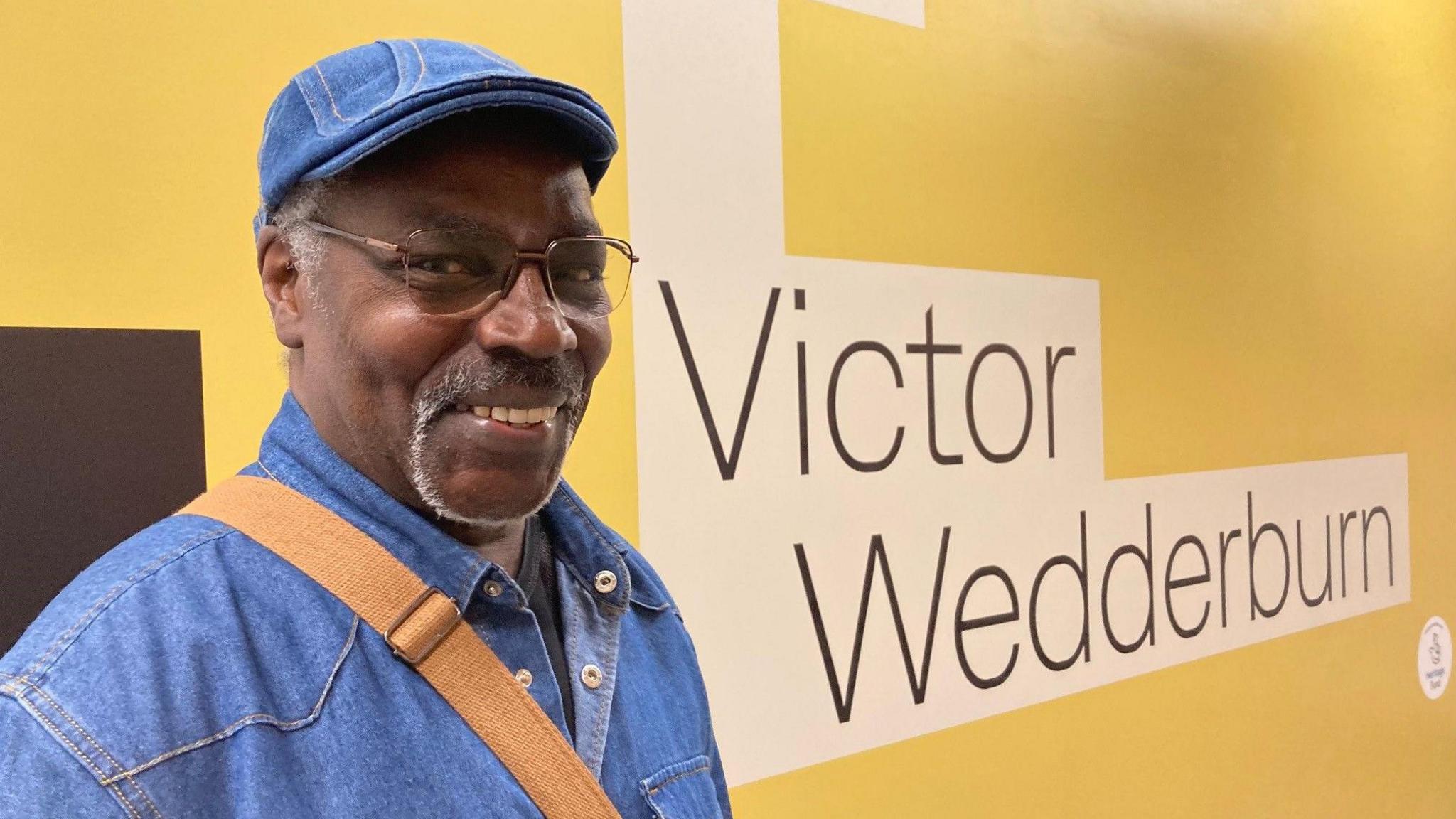 A man wearing a denim cap and shirt standing in front of wall.