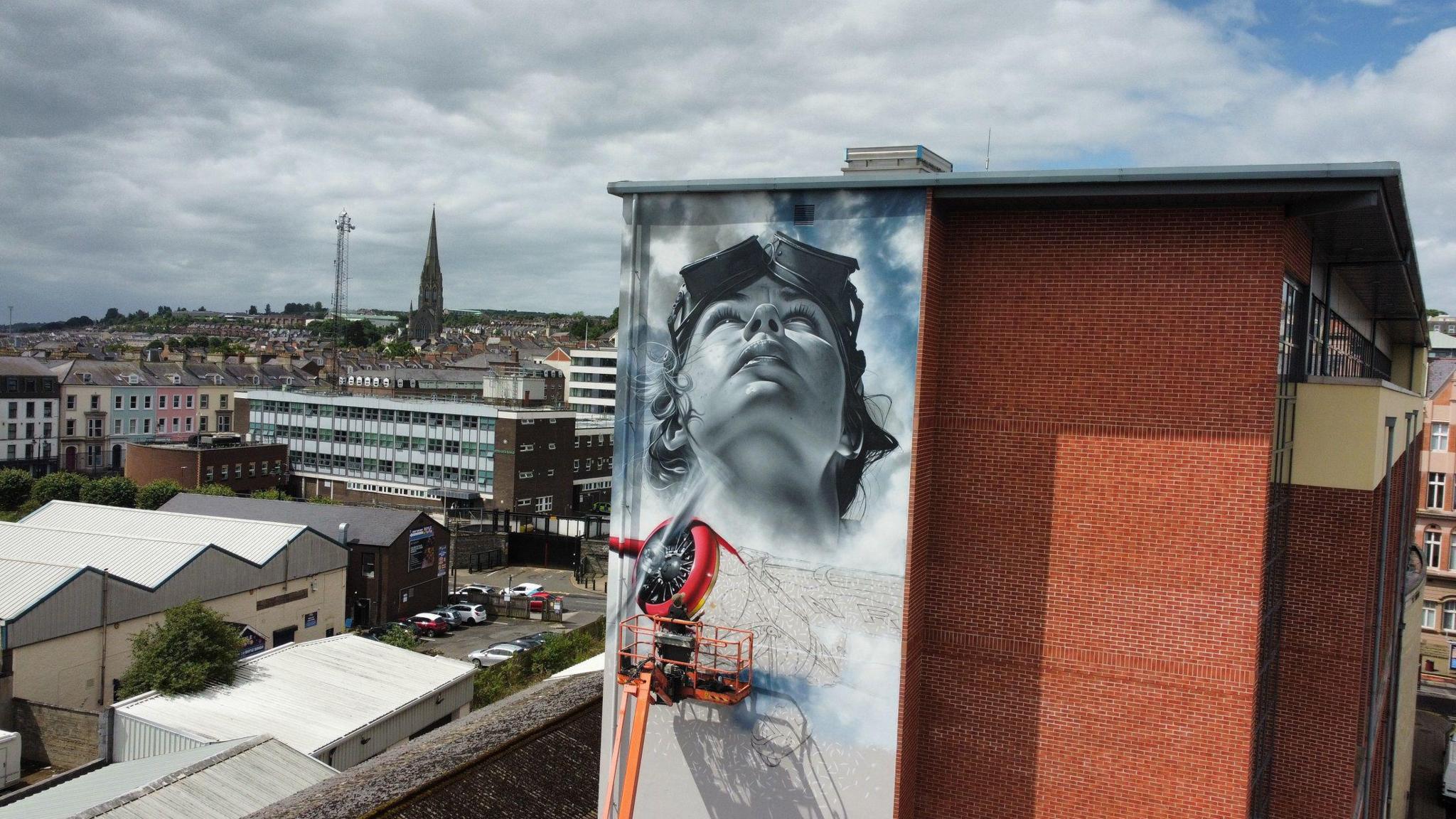 Artist jeks stands on a cherry picker while paiting a mural of Amelia Earhart on the Foyle building in Derry, in the backgournd are the city's rooftops