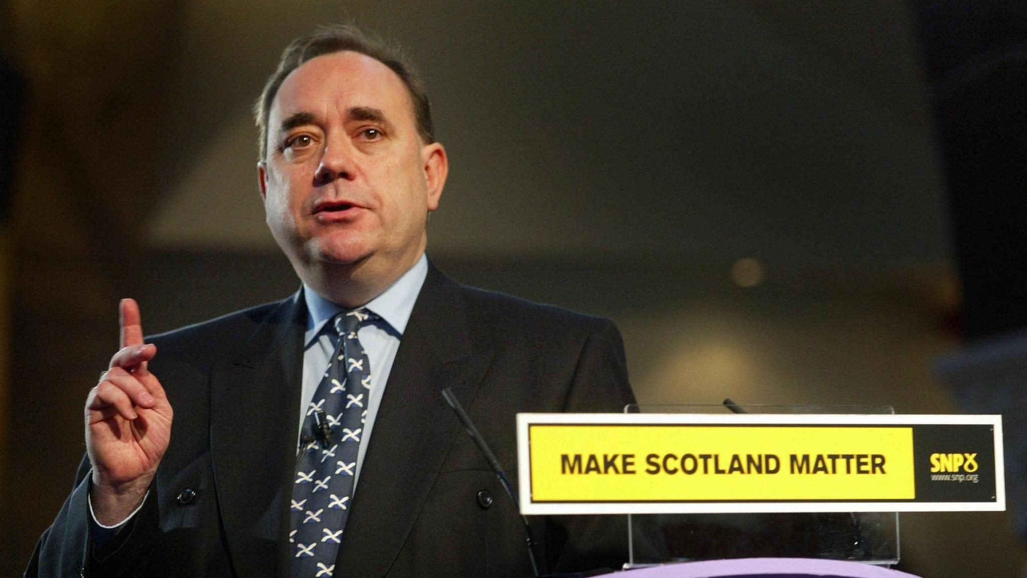 Alex Salmond holds a finger up while speaking, next to a sign saying "Make Scotland matter", wearing a dark suit, a light blue shirt and a dark tie with white crosses. He has brown hair. 