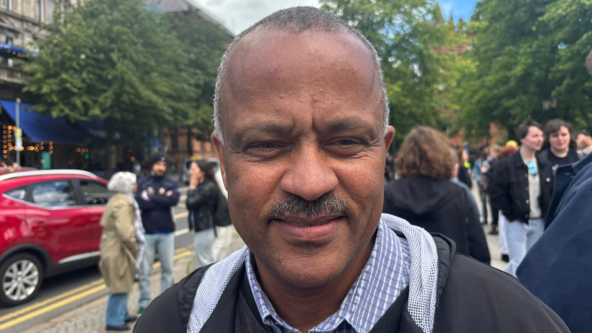 Mohamed is standing in front of a large group of people. He is wearing a black coat and blue shirt. He is smiling at the camera. 