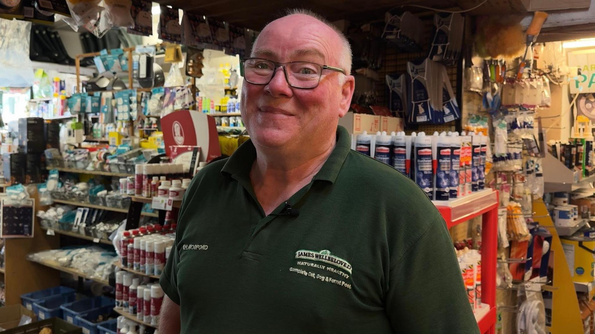 Paul Gillam is smiling at the camera inside his hardware shop. He is wearing a grey t-shirt and black framed glasses. The shelves are full of DIY supplies like paintbrushes, grout, paint and tape.