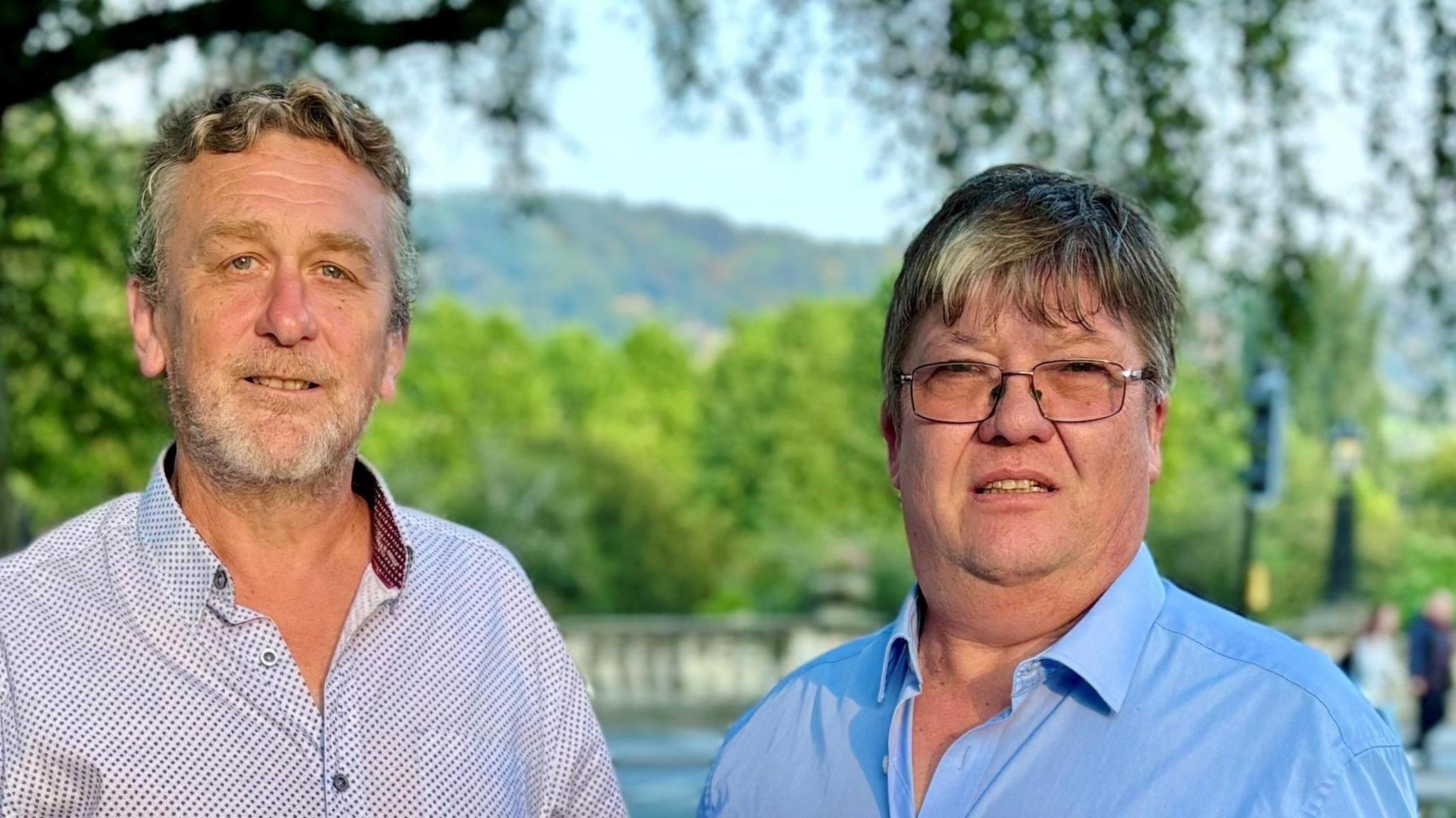 David Harding on the left wearing a white and blue patterned shirt, standing beside Michael Auton on the right, who is wearing a blue collared shirt and glasses. They are both standing in front of a body of water with trees visible in the background.