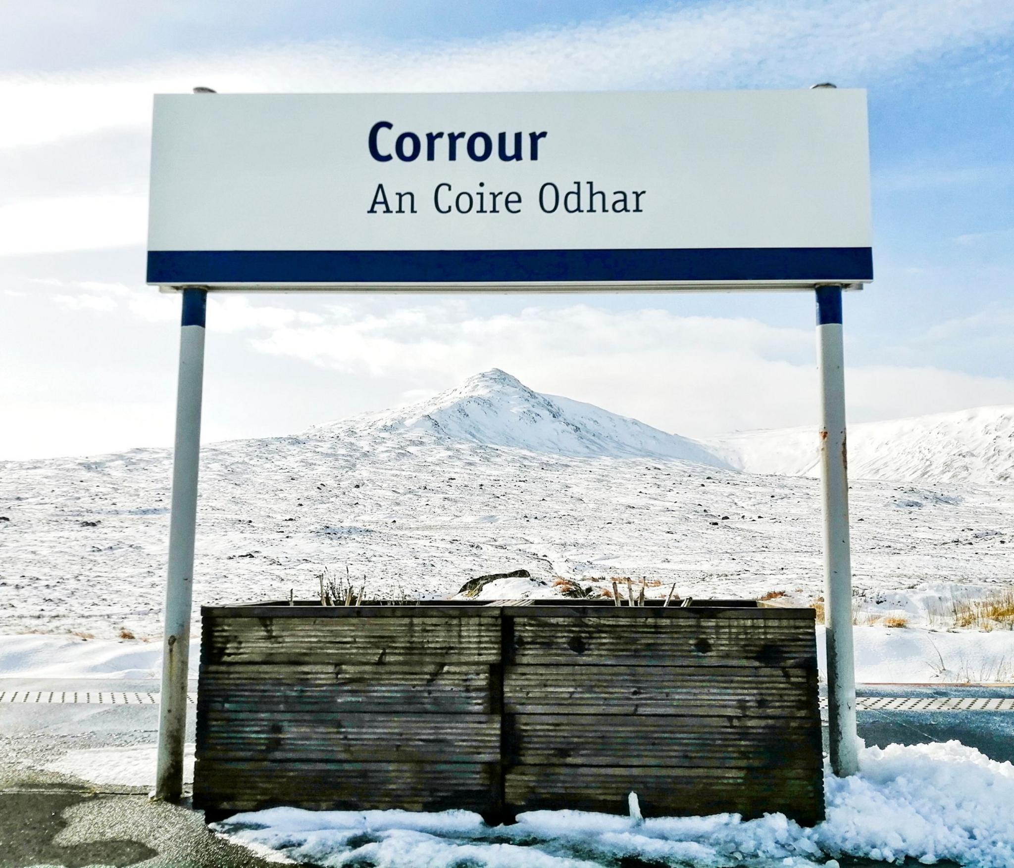 A white and blue sign at Corrour Station that reads the name Corrour in English and Gaelic. There is a mountain peak framed by poles holding the sign. There is snow on the mountain and the ground around the sign.