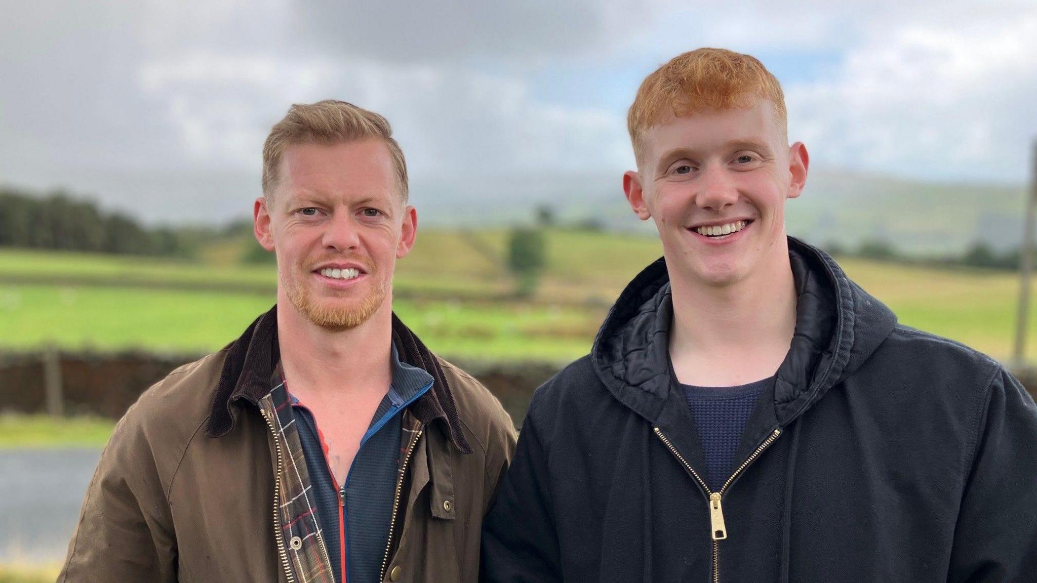 Two men stand next to each outdoors with green fields behind them. They are wearing coats and smiling.