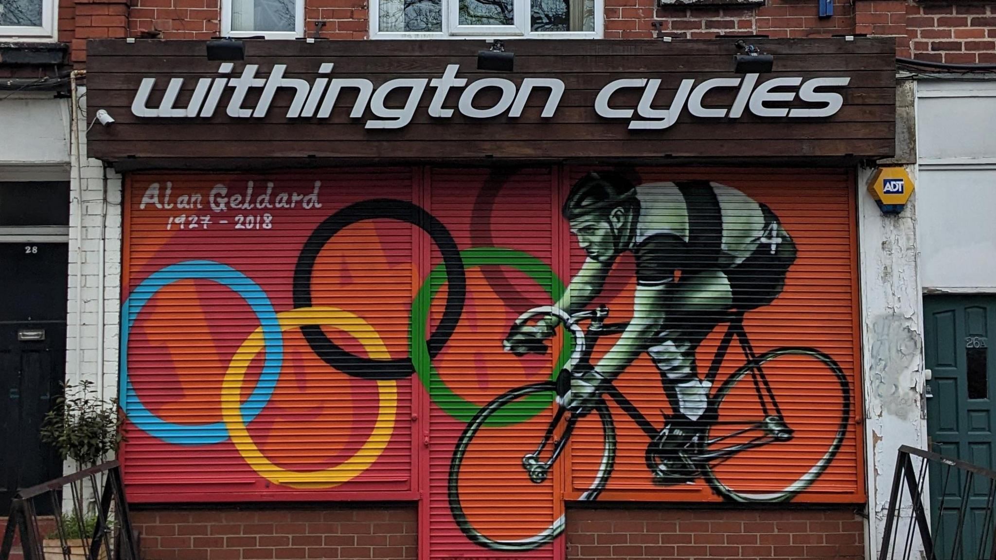 Alan Geldard on his bike aginst the Olympic rings painted on a shop shutter in Withington