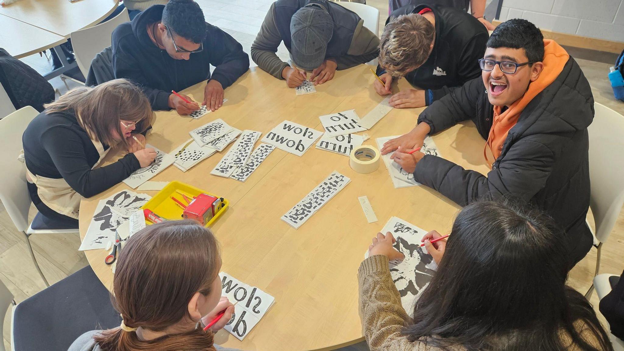 Seven teenagers sit around a round wooden table drawing and writing. One looks up at the camera grinning.