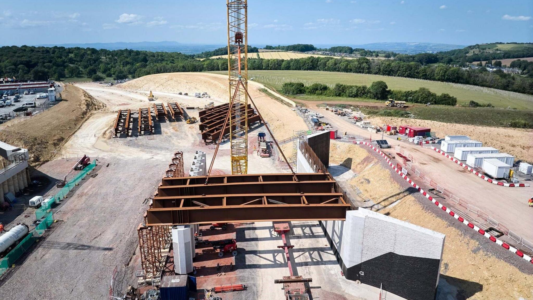 Another aerial shot of the work, this time from a lower angle taken at around crane height, rather than a birds-eye view. The shot shows a number of huge beams, one of which is suspended by straps from a yellow crane. Lots of vehicles and temporary structures can be seen nearby. 