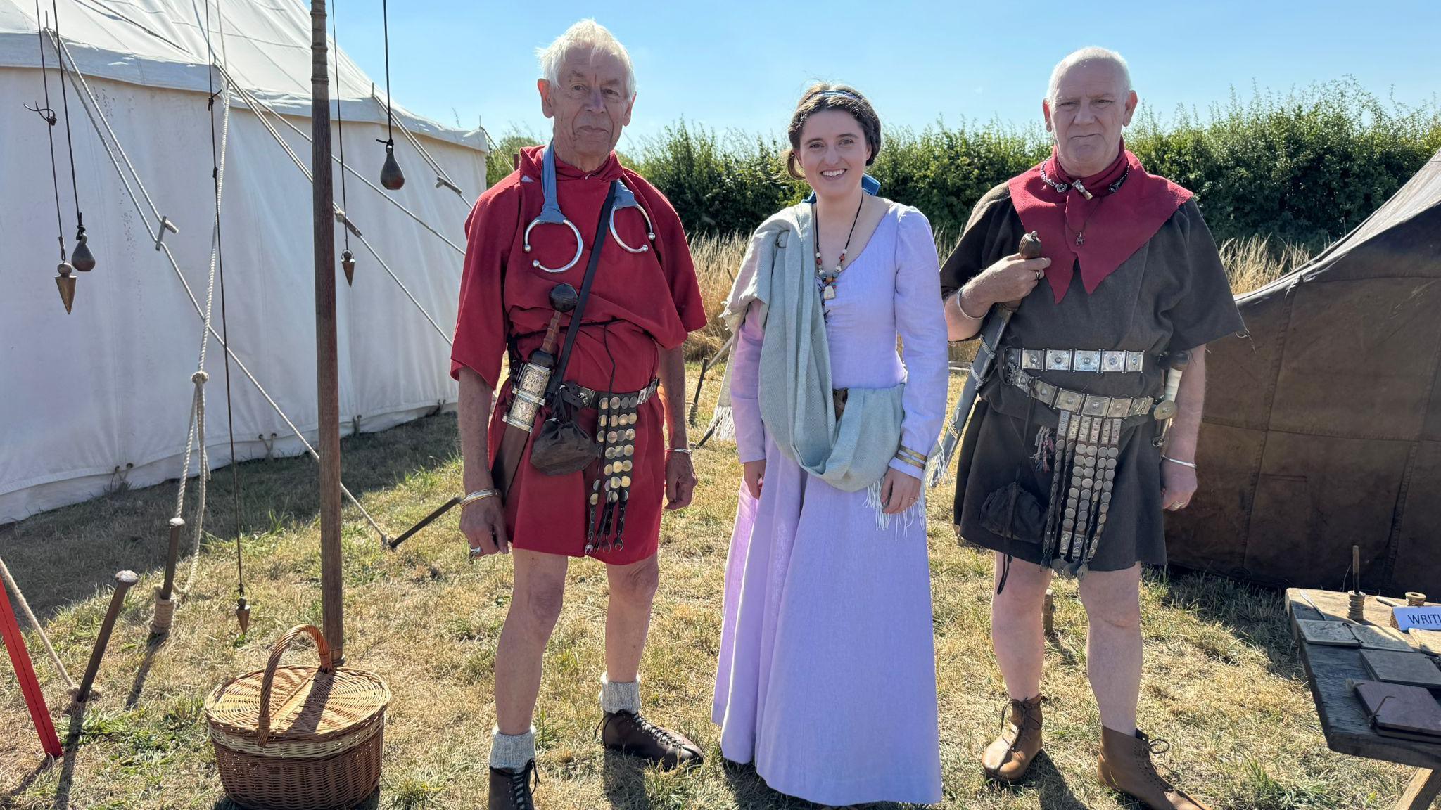 Three people pose next to a large white marquee in a field. Two men are dressed in red and brown Roman dress, while a woman in the middle wears a long pale blue dress and a light blue shawl. 
