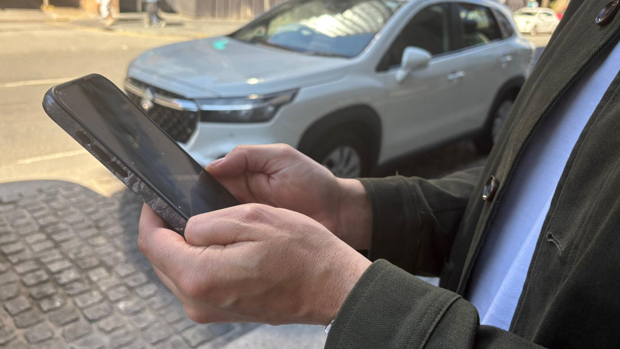 A man stands in a street holding up an Iphone. A car is parked behind him.