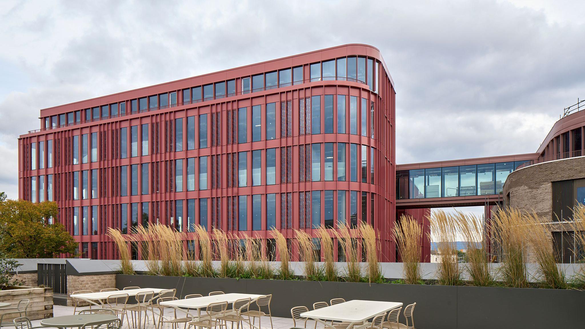The Forum in Gloucester viewed from a rooftop with chairs and tables. The Forum is a large, modern building with red coloured cladding and a glass walkway which connects it to another part of the building, which is brown brick.
