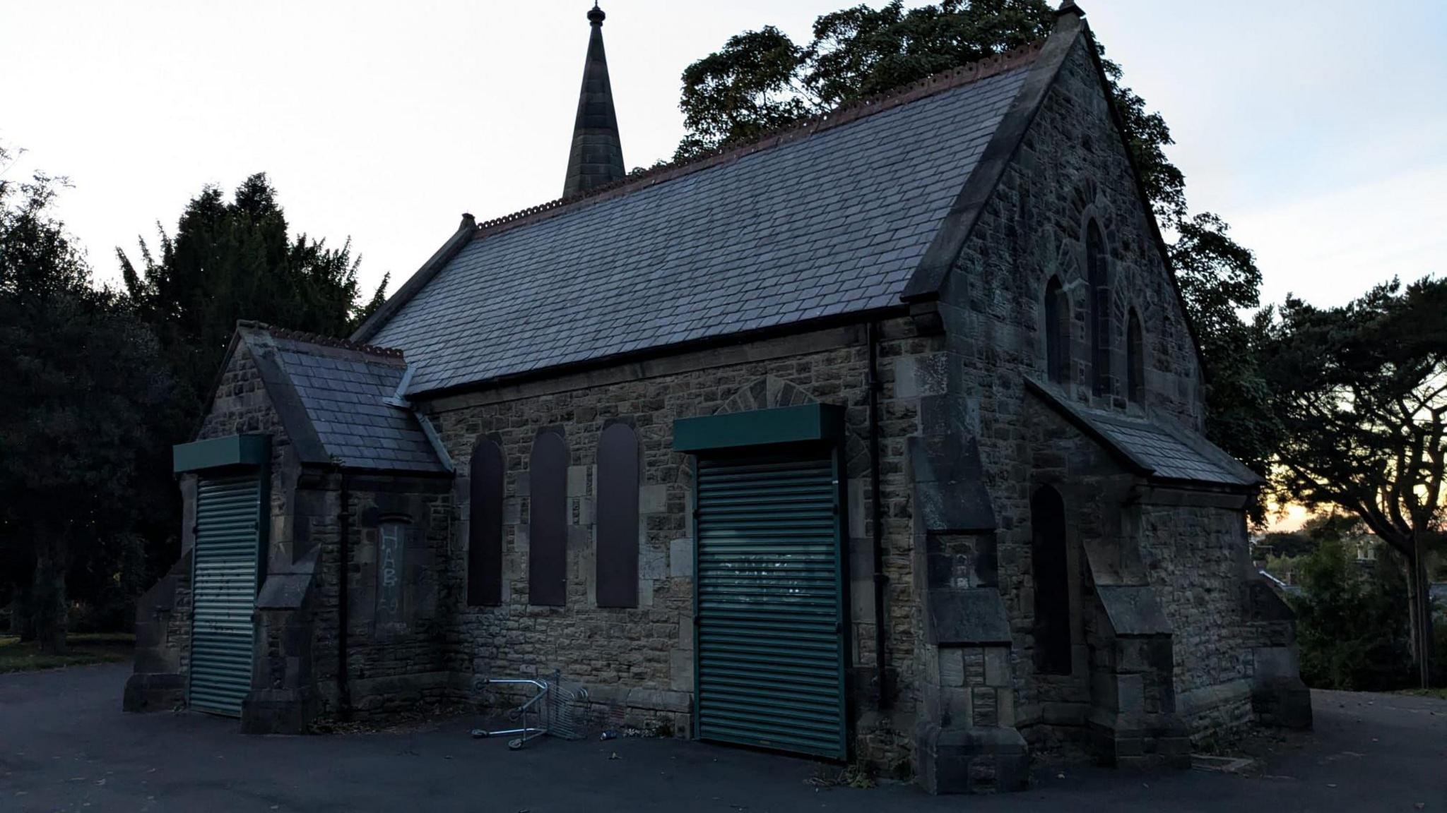 Ryton Chapel.  The stone chapel's windows have been boarded up and green shutters have been placed over its doors to bar entry. A shopping trolley lies overturned next to the building.