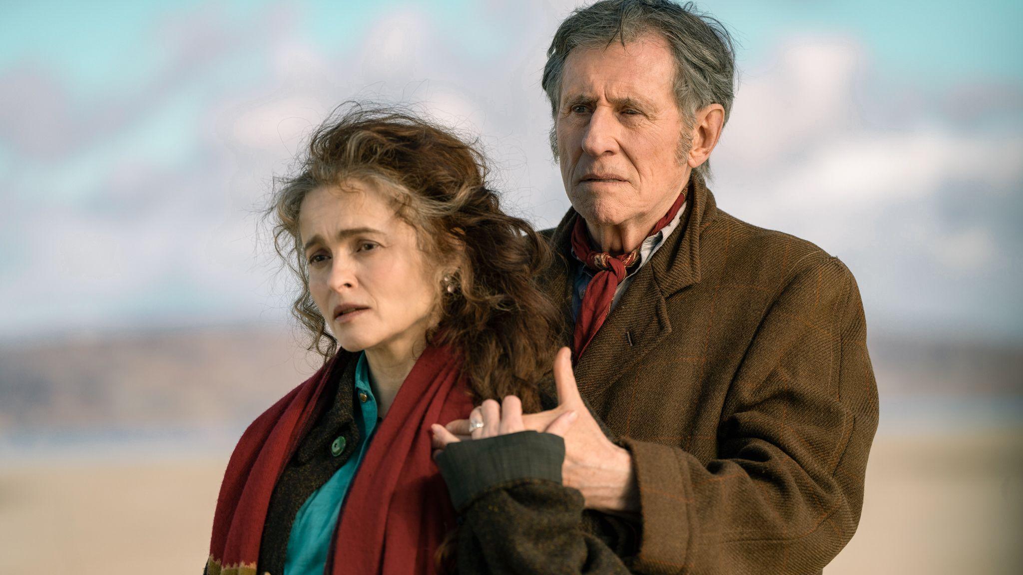 Helena Bonham Carter is wearing a red scarf and stands holding Gabriel Byrne's hand, he is dressed in a brown coat. They are on a beach.