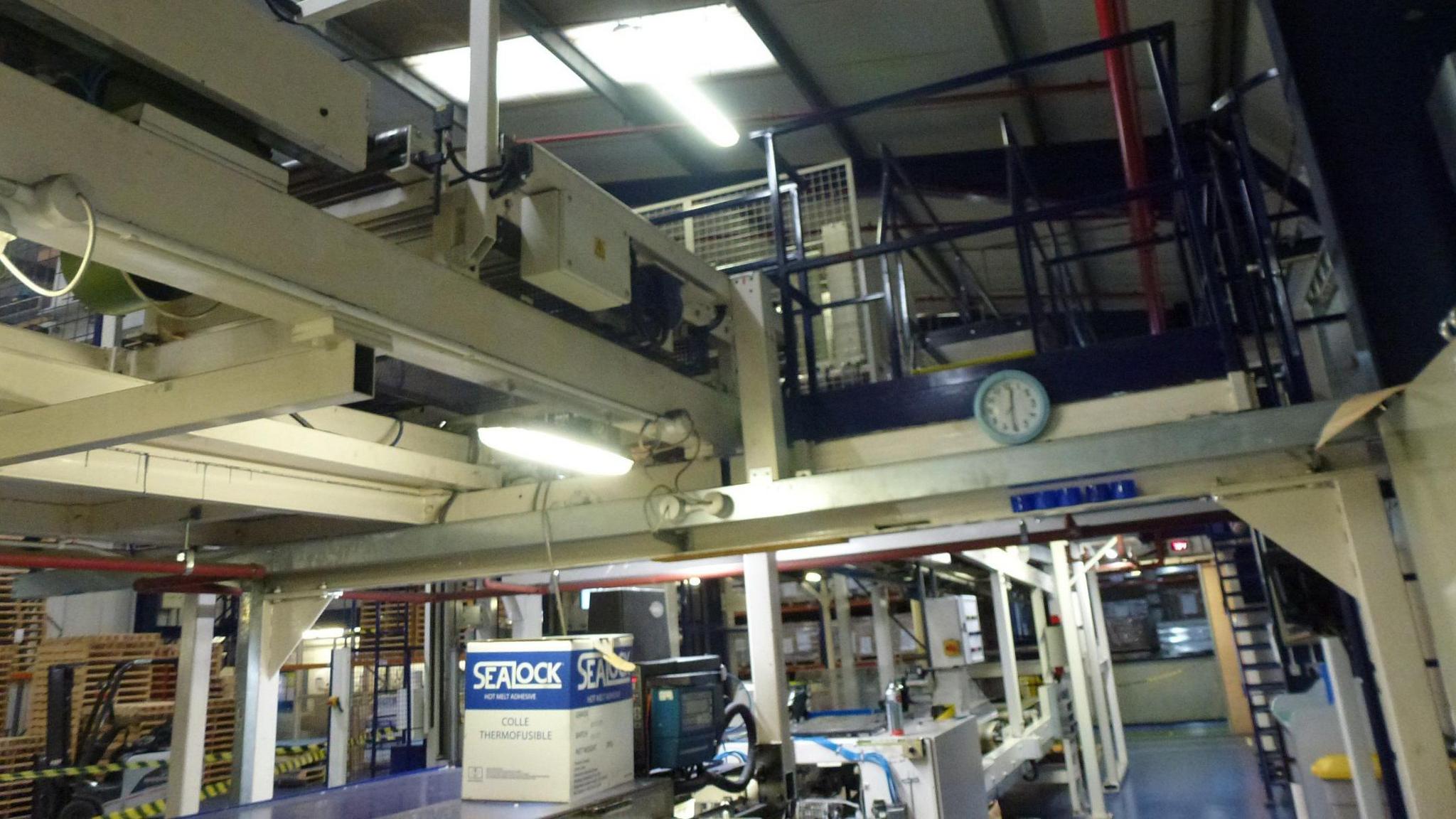 A factory floor with machines overhead. There is a clock attached to a metal bar in the distance.