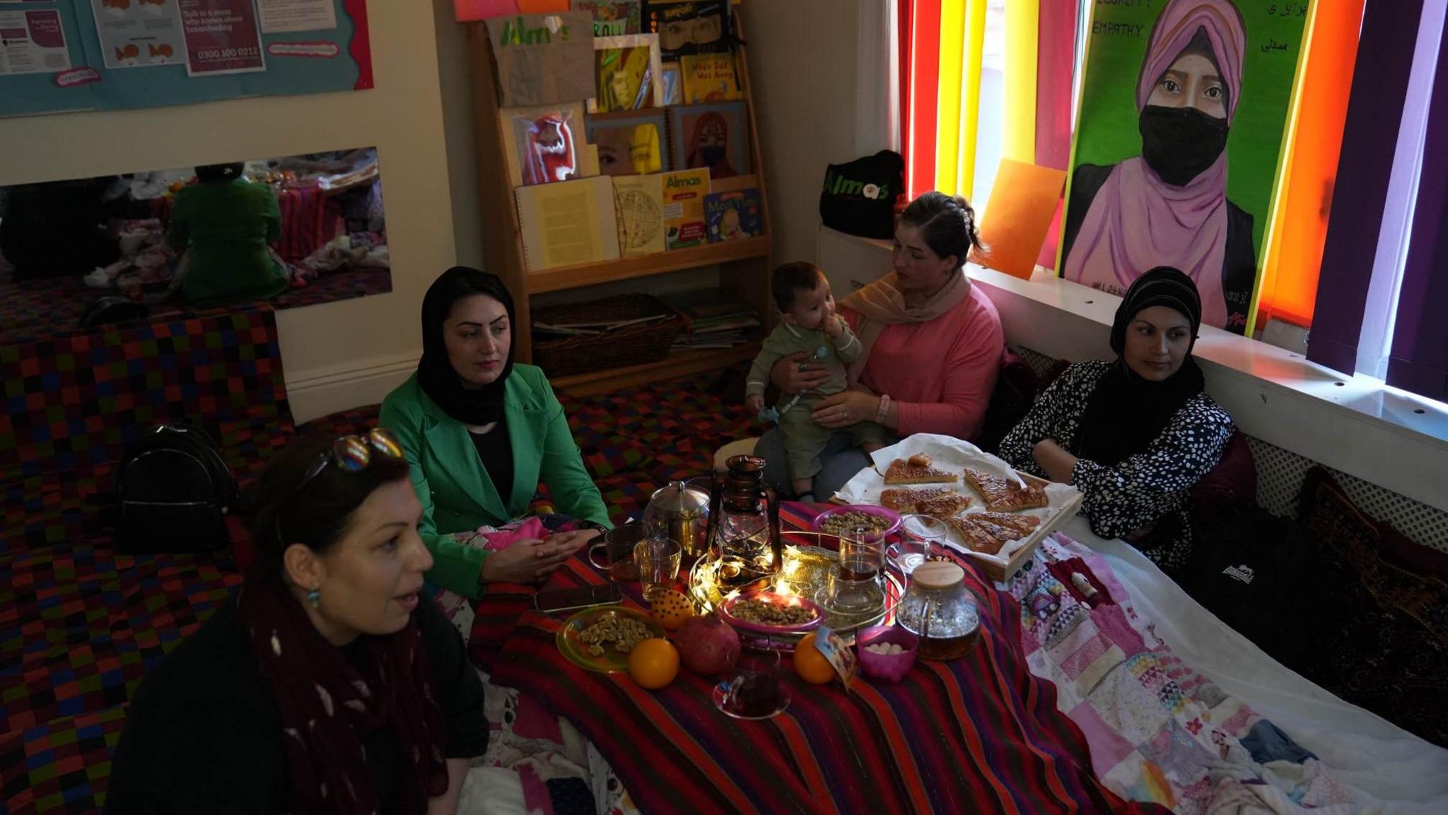 A group of women sitting around a table. Some women are wearing hijabs and there is tea and food on the table 