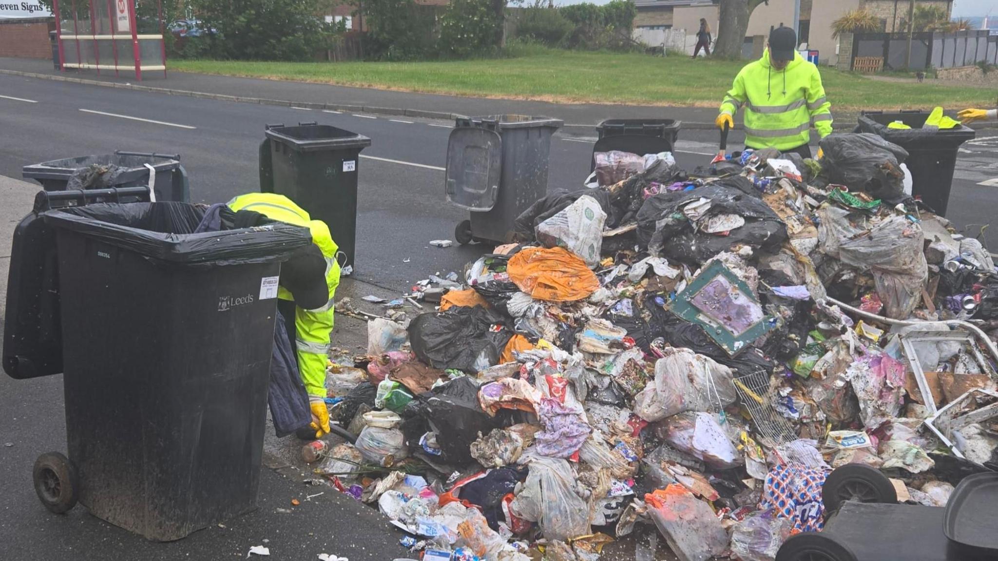 Two men in high vis jackets working on a large pile of rubbish, which has been emptied on to a road. Black wheelie bins surround the pile. In the background on the other side of the road is a bus stop and a pedestrian in the distance.