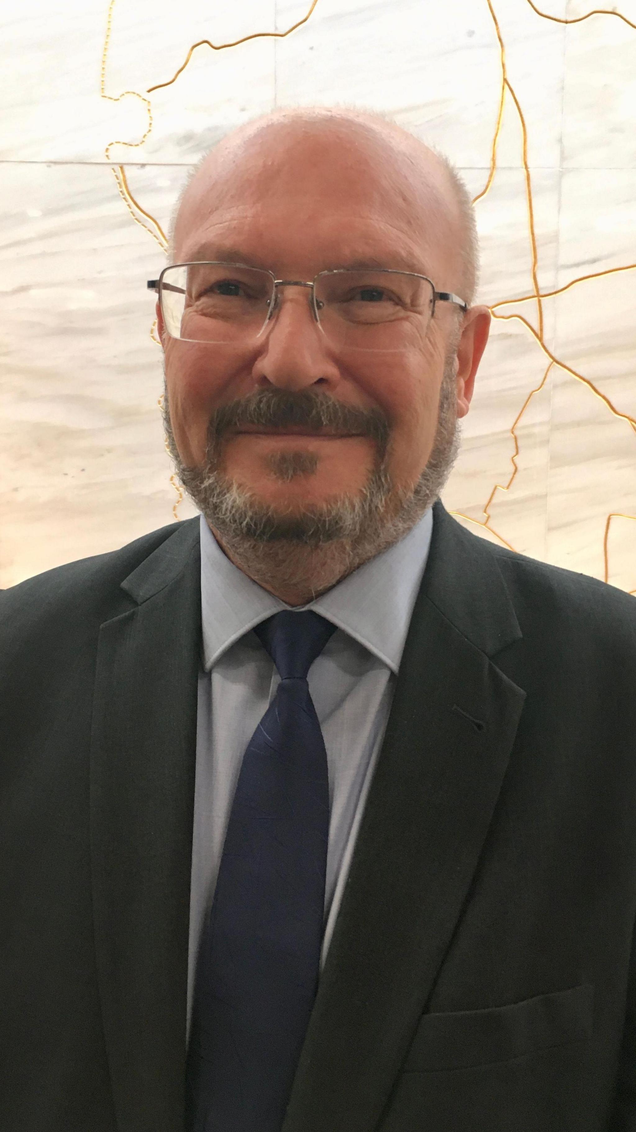 Graham Plant wearing a grey suit and a blue tie. He is bald, has a beard and wears silver-framed glasses