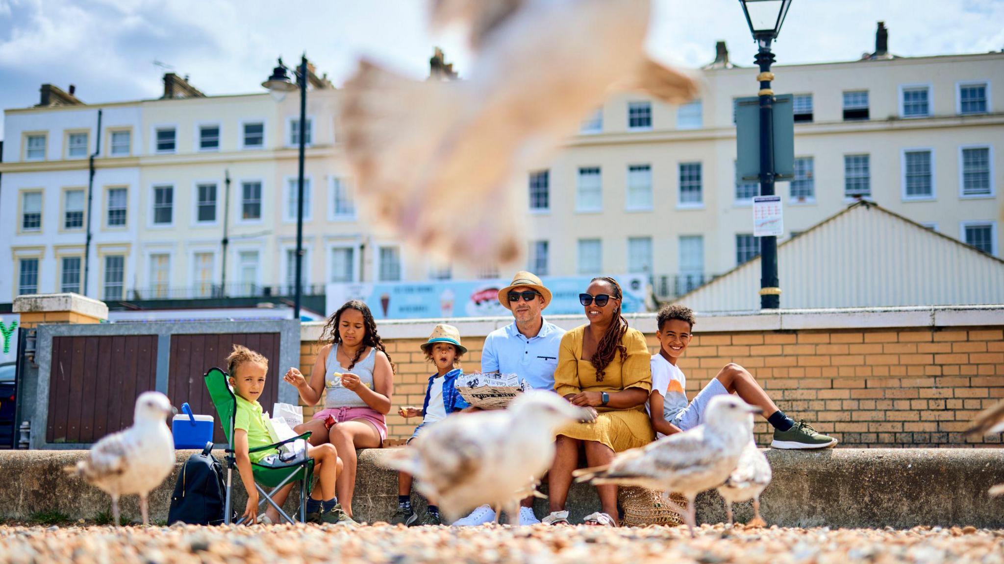 A seagull flies and several stand in front of a family sitting along a wall by a pebble covered beach at Herne Bay in Kent. A young boy is sitting in a green beach chair while a woman is sitting on the wall wearing a light grey top and pink shorts. Next to her is a boy with his mouth wide open who is wearing a trilby-style hat and blue over shirt with white t-shirt underneath. Then there is a man in a similar hat, sunglasses and a blue shirt, a woman in a yellow dress and another boy in t-shirt and shorts with his legs in a triangle along the wall.