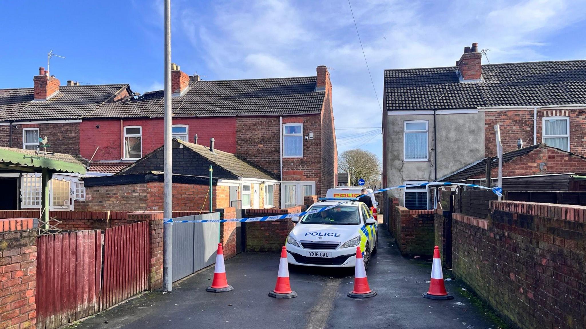 Teale Street in Scunthorpe. A street with residential houses on each side. A police car is parked in the middle of the street facing the camera. There is a blue and white police cordon and orange and white cones in front of the car.