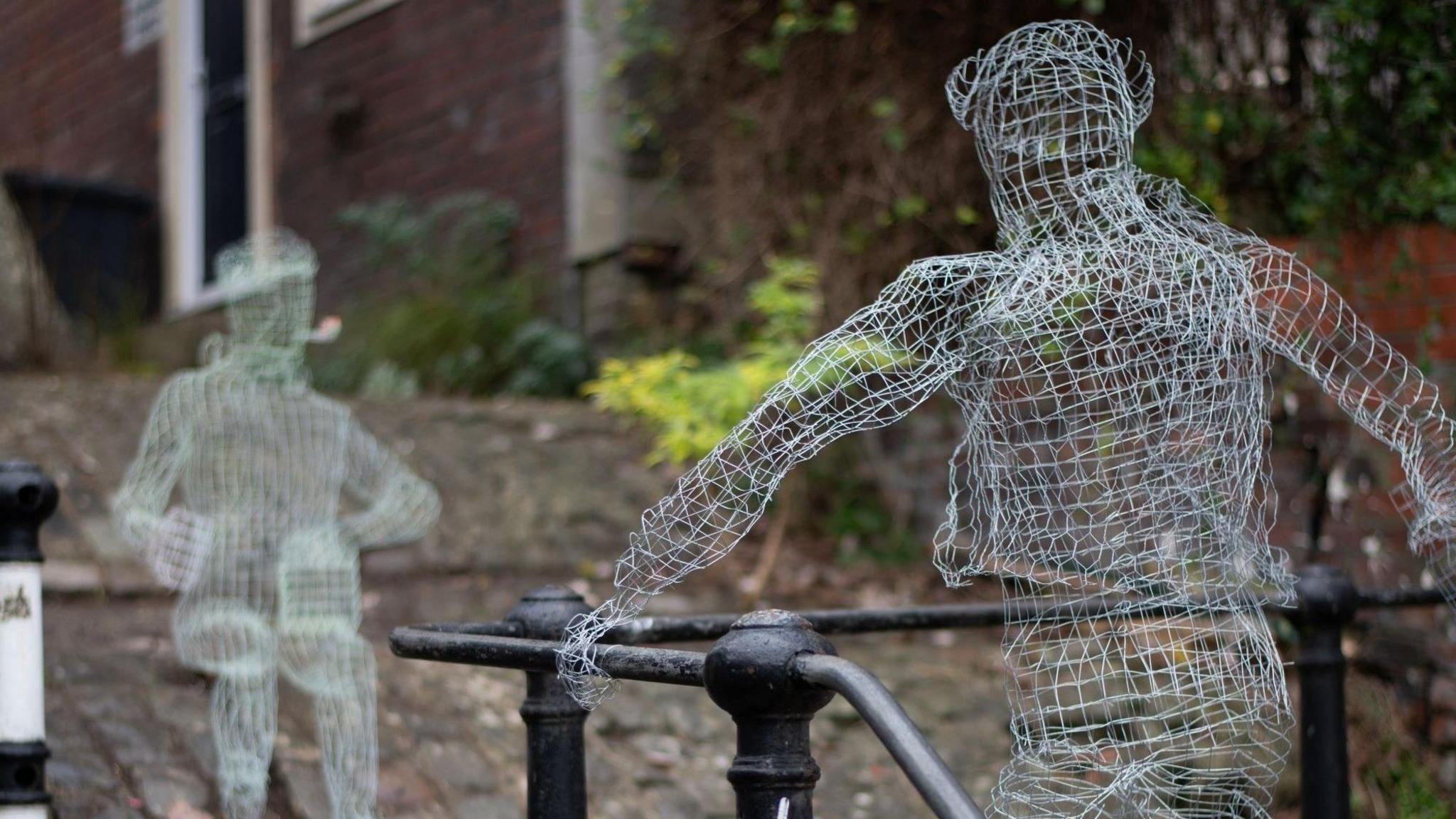 Two figures made from wire mesh face each other on the cobbles of St Luke's steps in Totterdown, Bristol. One of them is standing holding the rail alongside the stairs and another is sitting down smoking a pipe