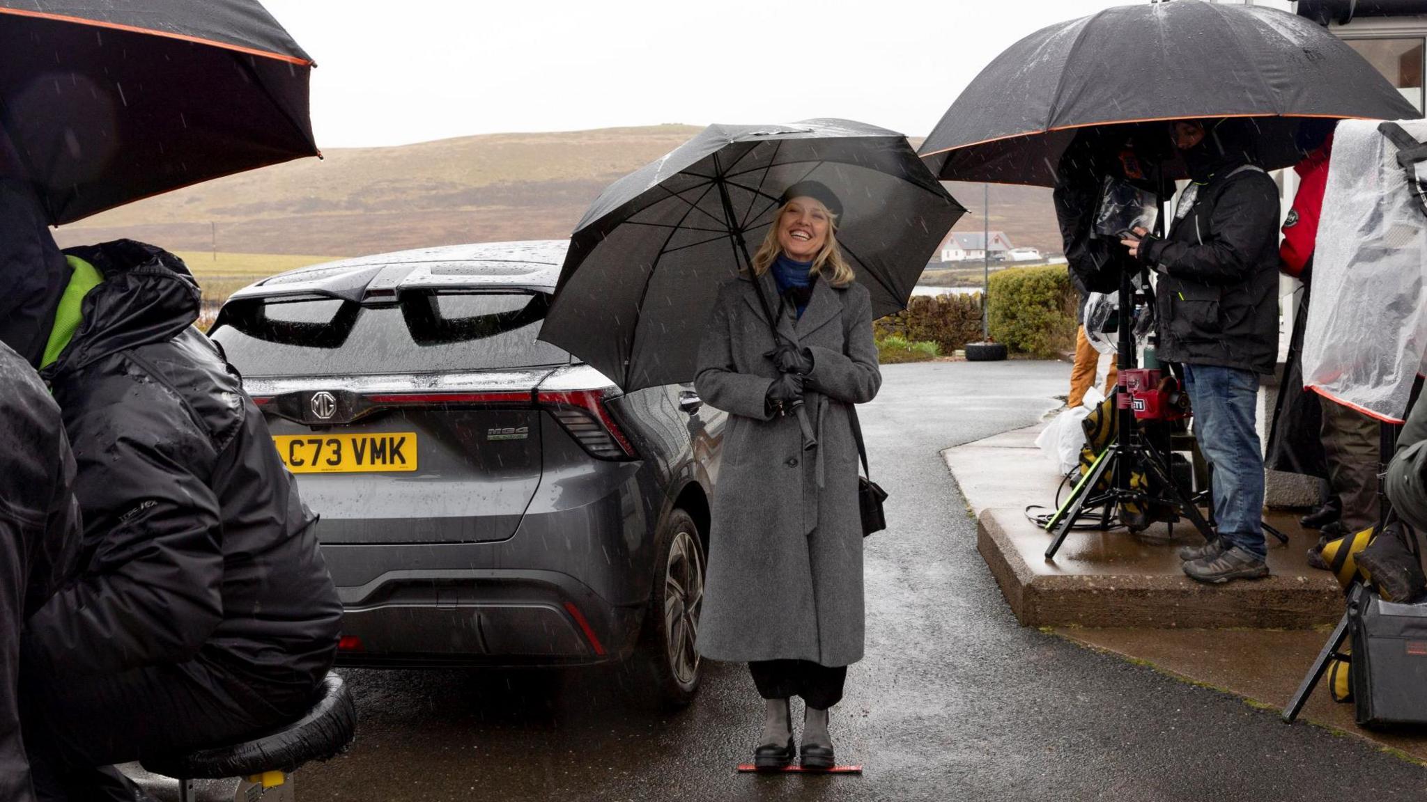 TV crew members work around Ashley Jensen, all with umbrellas and waterproof jackets on. Cameras have covers on to protect them from the rain. In the centre of the image, Jensen stands on a marker beside a car, a wet desolate hill behind her, but she is smiling at the camera holding a big golf umbrella, while being wrapped up in a long grey coat, a black beret and a blue and black scarf