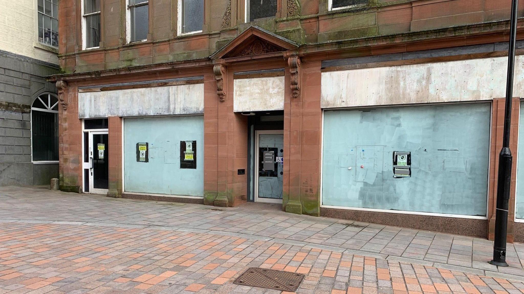 A boarded up shop front with posters in the window in a cobbled high street