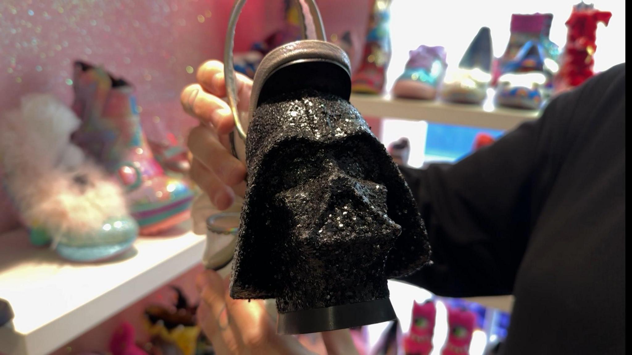 A high heeled shoe which has the Star Wars character Darth Vader's face mask as the heel. It's a glittery black colour