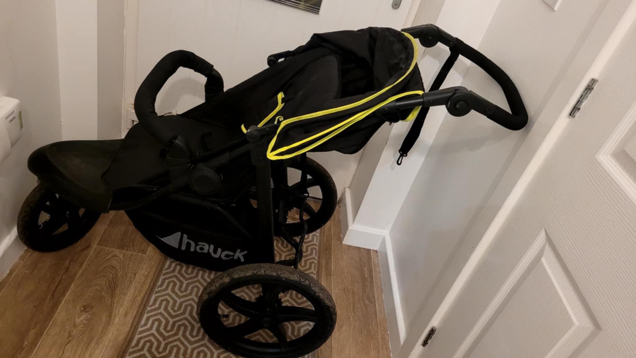 A black pushchair photographed in front of a front door with a runner carpet on the floor.