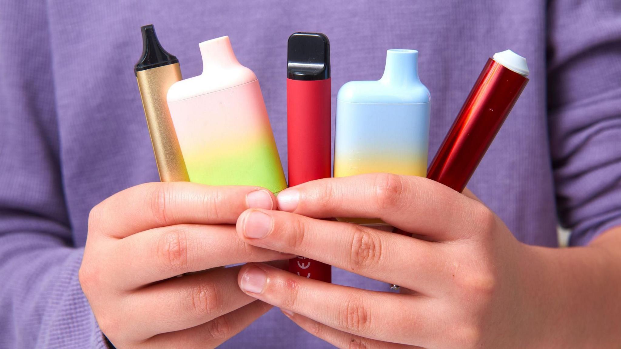 Someone wearing a lilac top holds five different coloured vapes. 