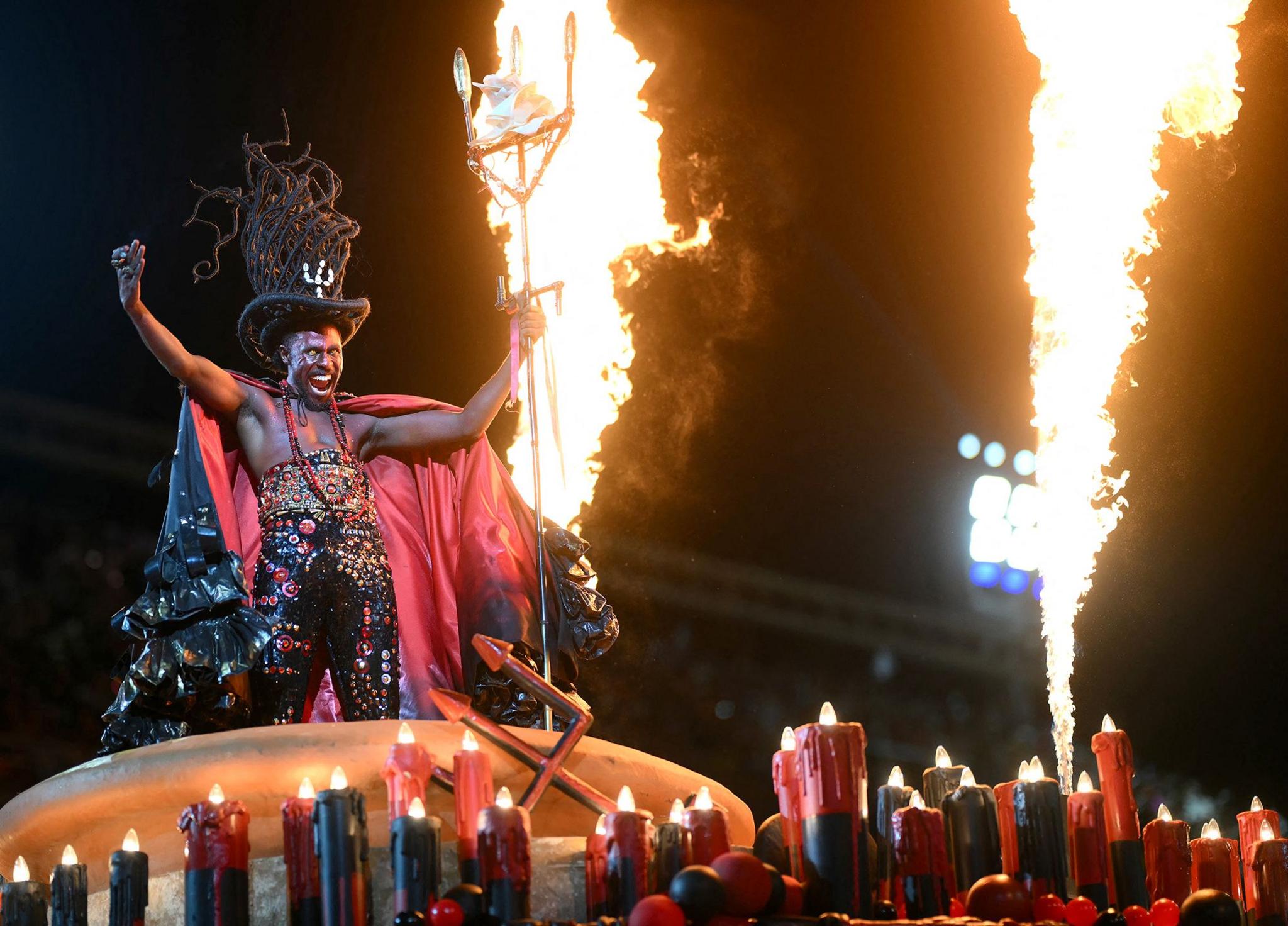 A reveler of the Beija-Flor samba school performs dressed as a devil. He is is gesturing and is surrounded by jets of flame. 