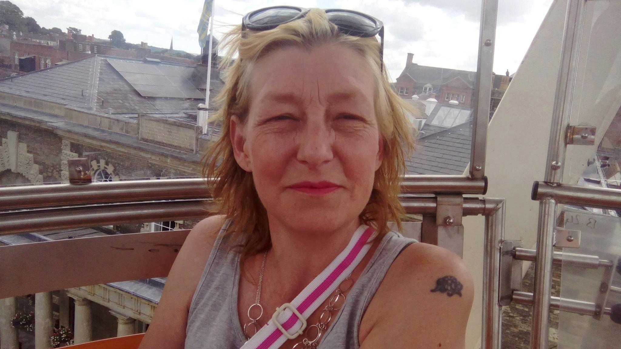 an image of a woman, sat on a rooftop with buildings behind her. She has light brown hair, is wearing a grey top and has a bag strap over her shoulder.