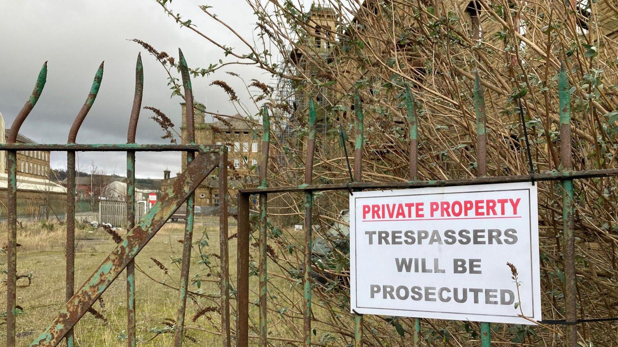 A bent metal sign attached to green, rusting railings which reads 'Private Property' in red lettering and then 'Trespassers will be prosecuted' in black underneath with part of the mill complex visible in the distance.