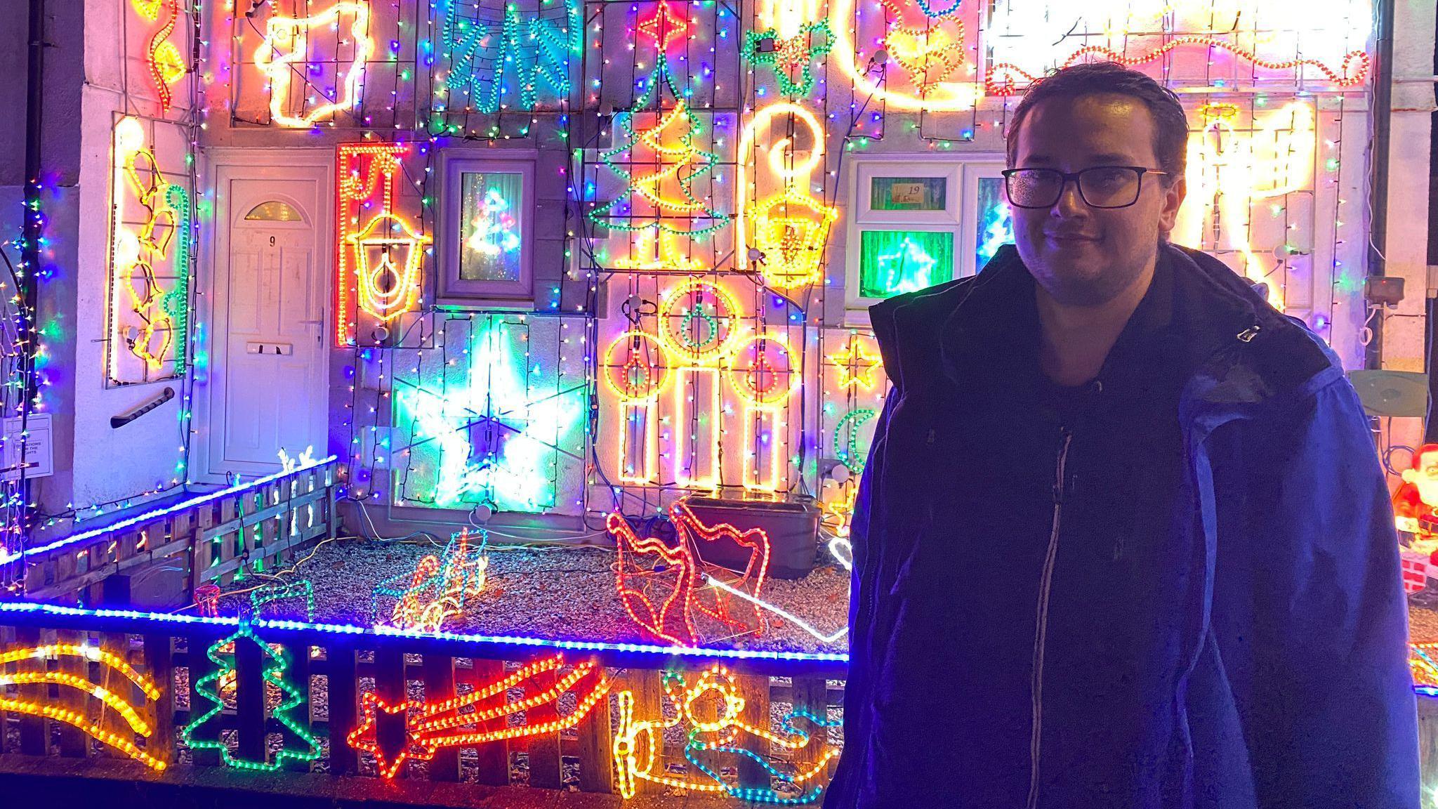 Dan Noall stands outside of the display in Cornwall. Mr Noall is wearing a blue coat on top of a jumper. He has dark hair and is wearing glases. Red, green, blue and yellow Christmas lights light up the background behind him.