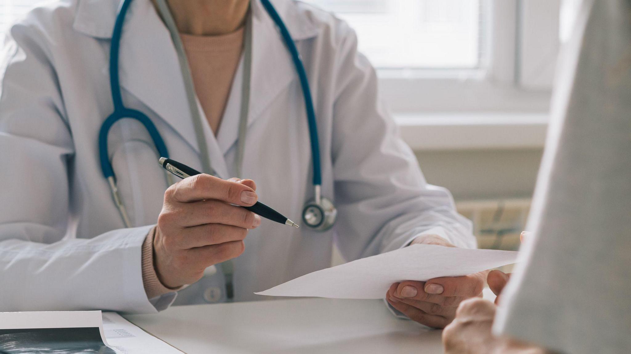 A doctor, wearing a white jacket with a stethoscope around their next and holding a pen and piece of paper. They are sitting at a desk, across from a patient, who is out of shot.  