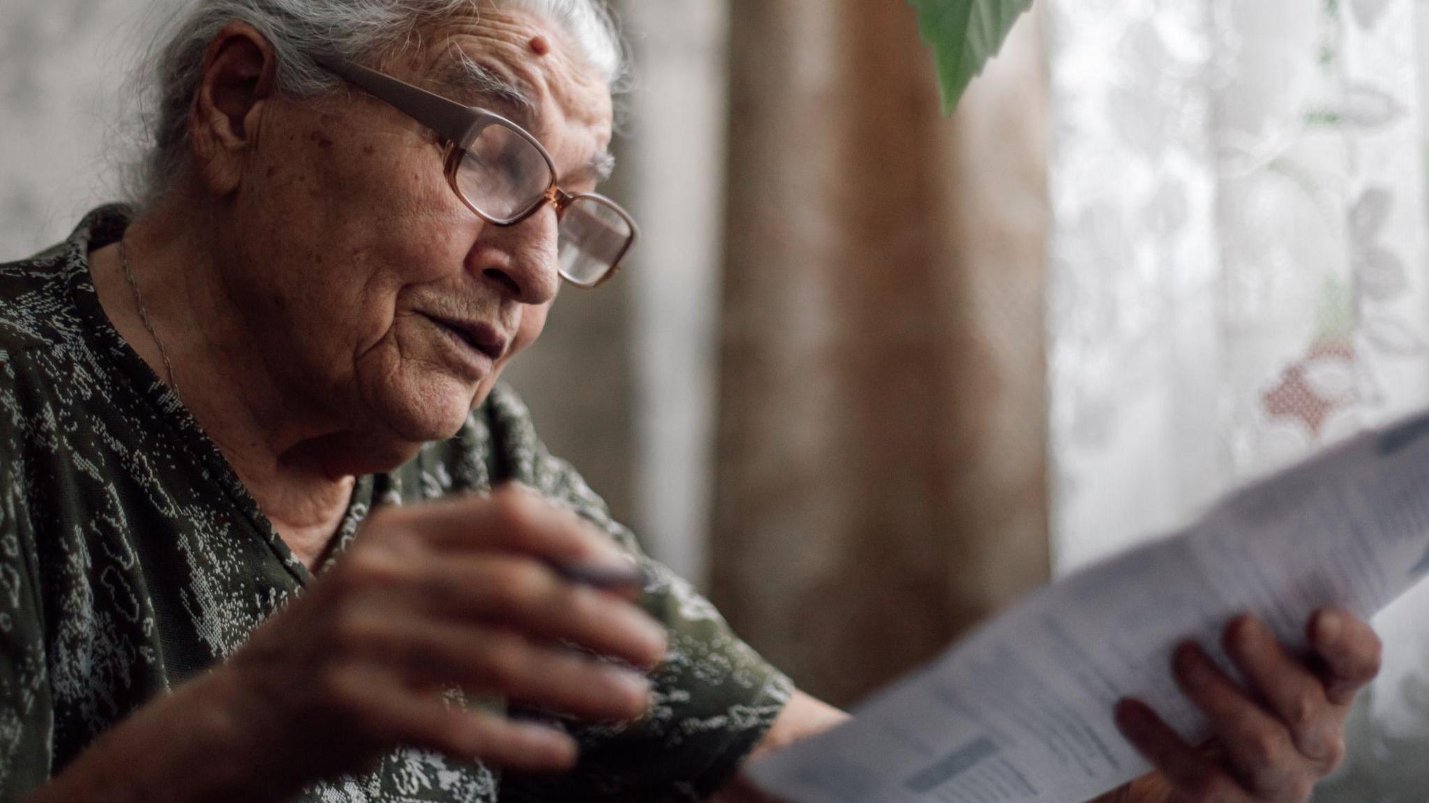 Older woman wearing glasses looks at a bill with curtains and net curtains behind her.